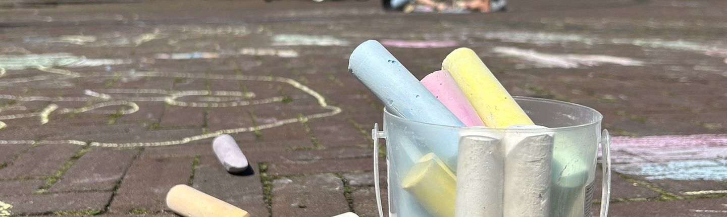 Becher voller bunter Kreide auf bemalten Marktplatz. Im Hintergrund malen Kinder.