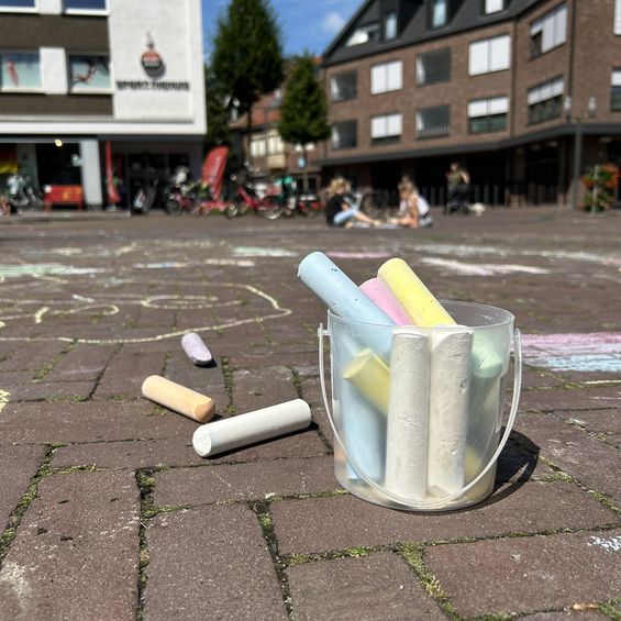 Becher voller bunter Kreide auf bemalten Marktplatz. Im Hintergrund malen Kinder.
