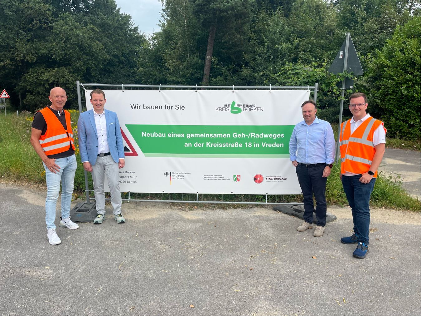 Es stehen vor einem Baustellenzaun: v. l. n. r.: Christian Abbing (Kreisbetrieb), Vredens Bürgermeister Dr. Tom Tenostendarp, Landrat Dr. Kai Zwicker und Alexandru Dan (Kreisbetrieb)