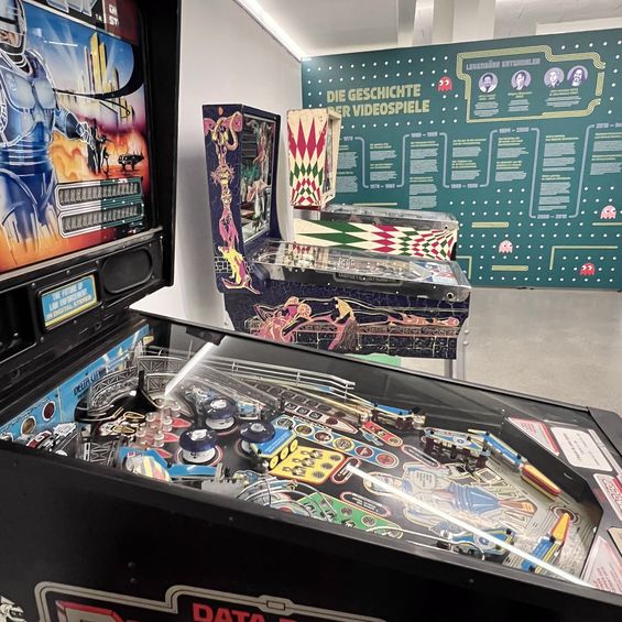 Bild einiger alter Spielautomaten mit einer Infotafel "Die Geschichte der Videospiele" im Hintergrund