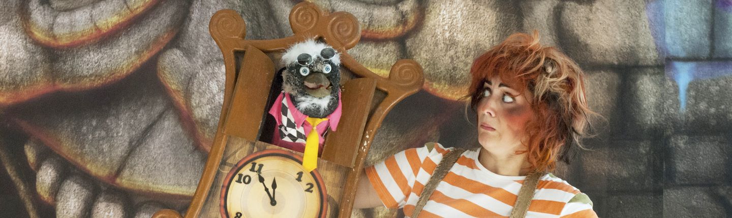 Aufnahme aus dem Theaterstück "Der kleine Zahlenteufel. Der Zahlenteufel schaut aus einer Kuckucksuhr. Eine Frau in Orange-weiß gestreiftem T-Shirt schaut ihn an.