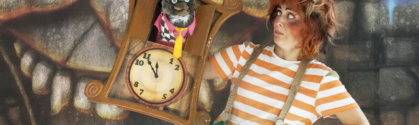 Aufnahme aus dem Theaterstück "Der kleine Zahlenteufel. Der Zahlenteufel schaut aus einer Kuckucksuhr. Eine Frau in Orange-weiß gestreiftem T-Shirt schaut ihn an.