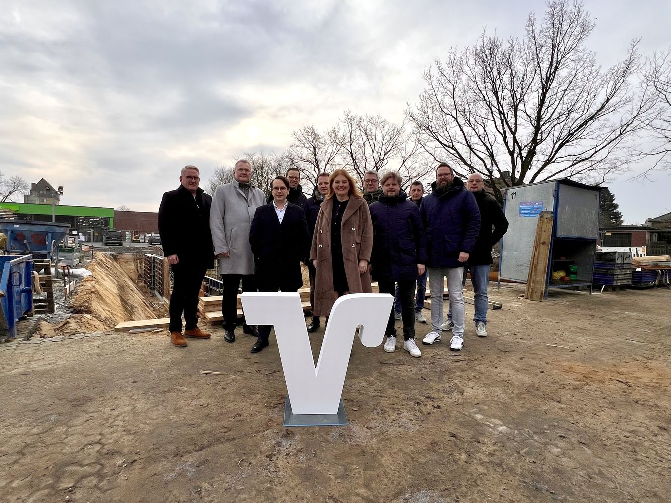 Gruppenbild der Beteiligten vor der Baustelle. Volksbank "V" im Vordergrund. Auf dem Foto sind zu sehen v.l.n.r.: Heiner Temmink, Georg Kremerskothen, Tobias Kühn, Benedikt Bengfort, Dr. Tom Tenostendarp, Simone Goertz, Vertreter der Firma Weitkamp + Partner mbB und Heinrich Temmink GmbH & Co. KG