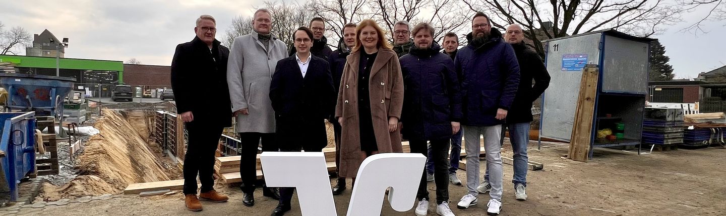 Gruppenbild der Beteiligten vor der Baustelle. Volksbank "V" im Vordergrund. Auf dem Foto sind zu sehen v.l.n.r.: Heiner Temmink, Georg Kremerskothen, Tobias Kühn, Benedikt Bengfort, Dr. Tom Tenostendarp, Simone Goertz, Vertreter der Firma Weitkamp + Partner mbB und Heinrich Temmink GmbH & Co. KG