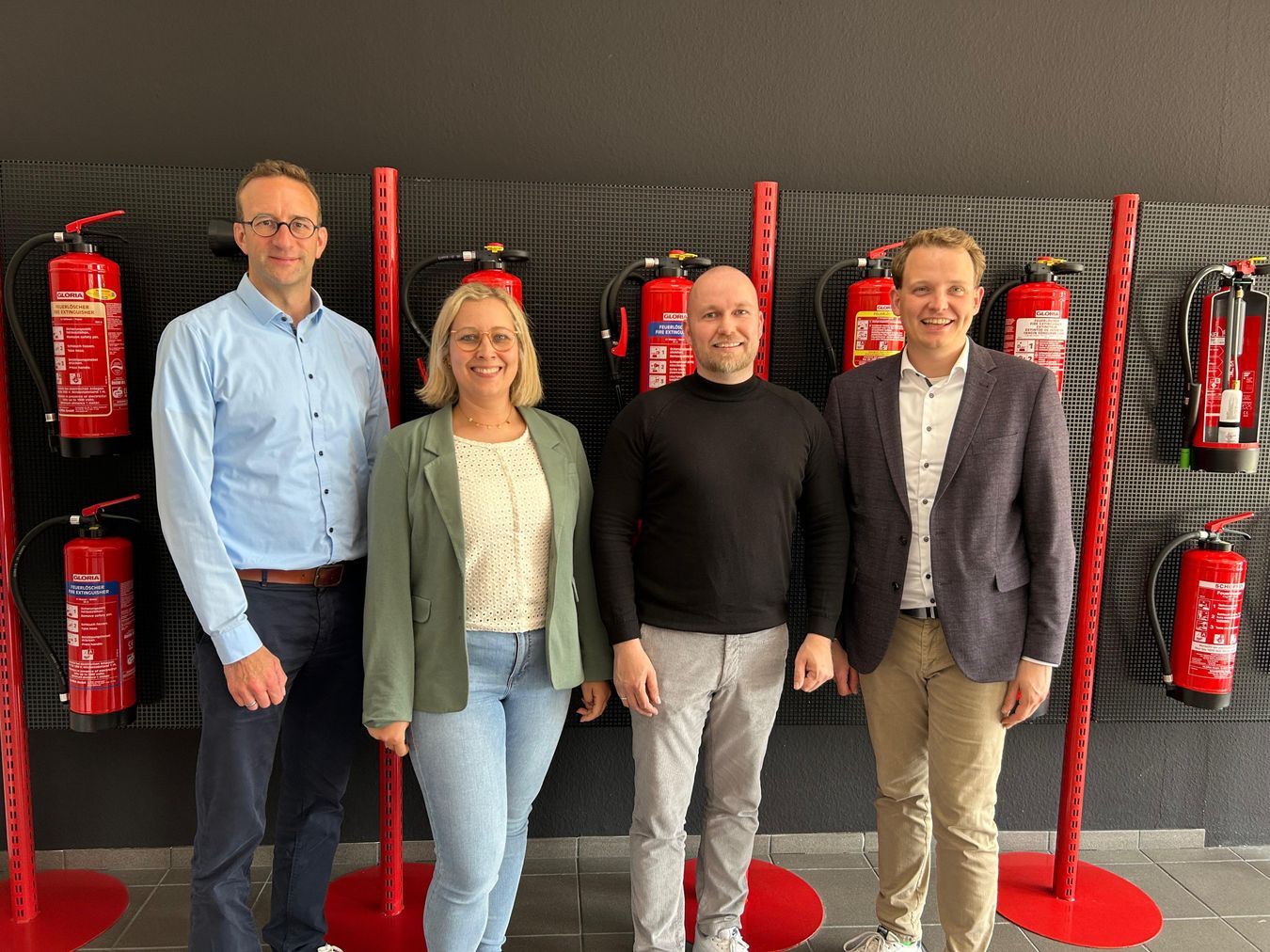 Jana und Henning J. Schoppen von Brandschutztechnik Schoppen (Mitte) sowie Wirtschaftsförderer Michael Terhörst (links) und Bürgermeister Dr. Tom Tenostendarp (rechts) vor einer Wand voller Feuerlöschern.