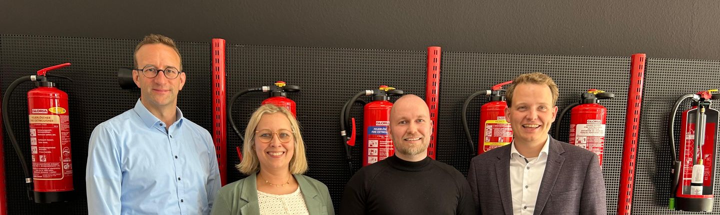 Jana und Henning J. Schoppen von Brandschutztechnik Schoppen (Mitte) sowie Wirtschaftsförderer Michael Terhörst (links) und Bürgermeister Dr. Tom Tenostendarp (rechts) vor einer Wand voller Feuerlöschern.