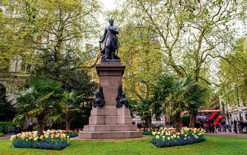 Garden close to the River Thames Statue in a park surrounded by green trees, palm plants, and colourful flowers, with a red double-decker bus in the background.