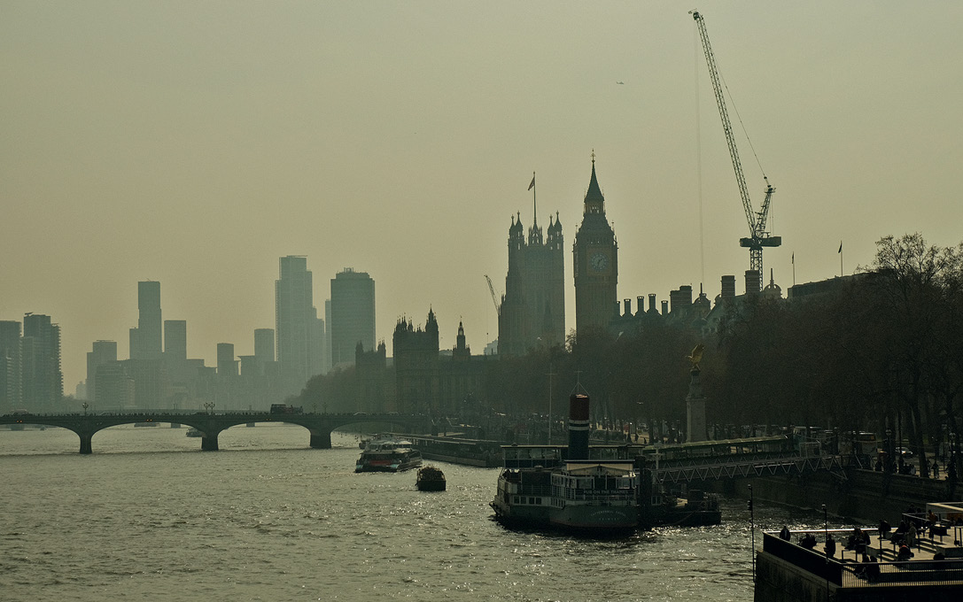 Silhouette of London's iconic skyline with Big Ben, cranes, and skyscrapers under a hazy sky, viewed from the River Thames.