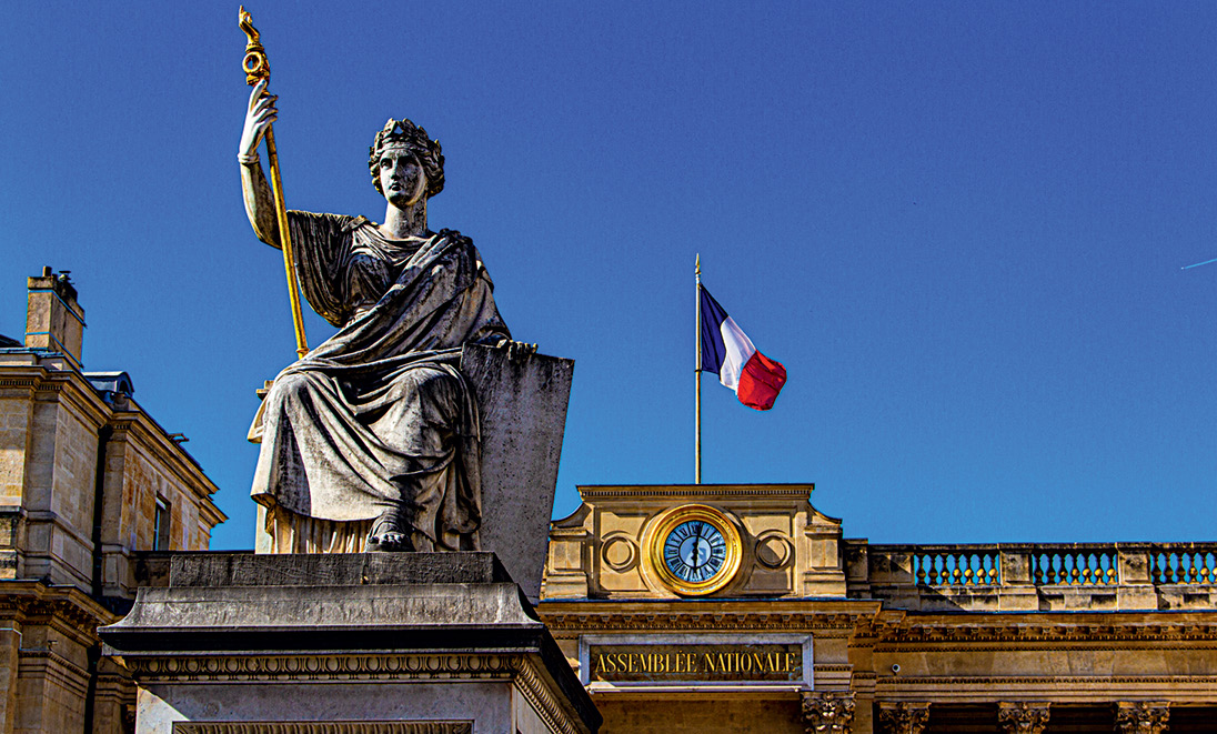 A statue in front of the National Assembly in Paris. Statue holding a staff in front of the French National Assembly building, with the French flag and a clock on the façade.