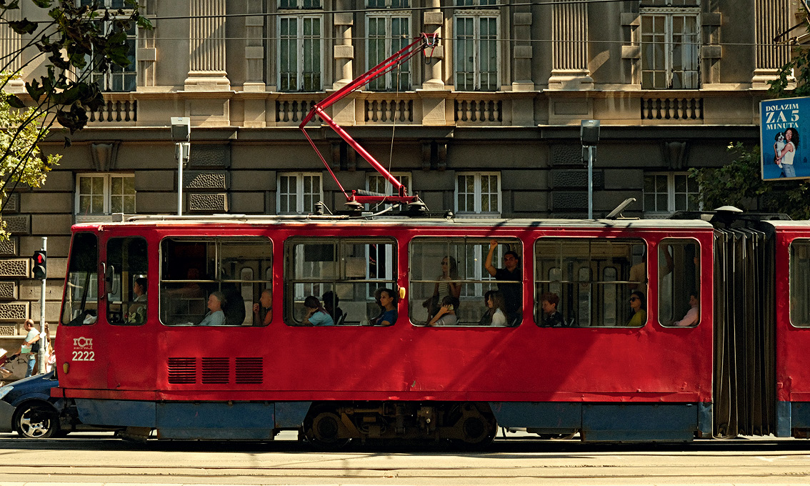 Red tram with passengers travels along a city street lined with an old building featuring large white-framed windows.