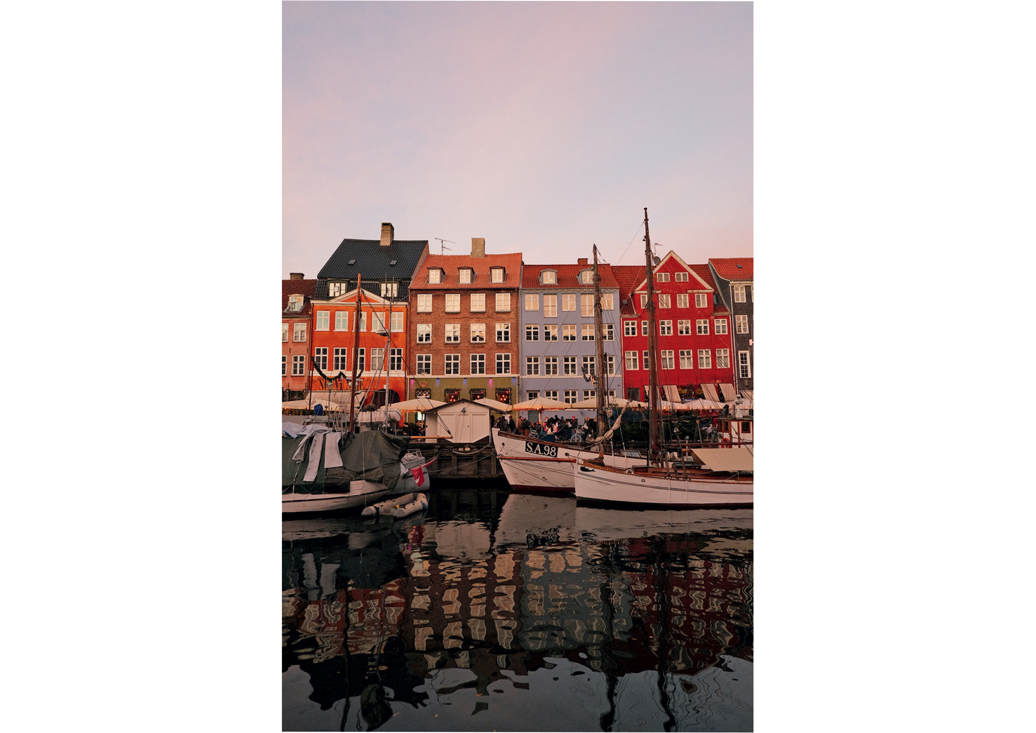 Colorful buildings line a waterfront with boats in the harbor, reflecting in calm water under a pink-tinged sky.