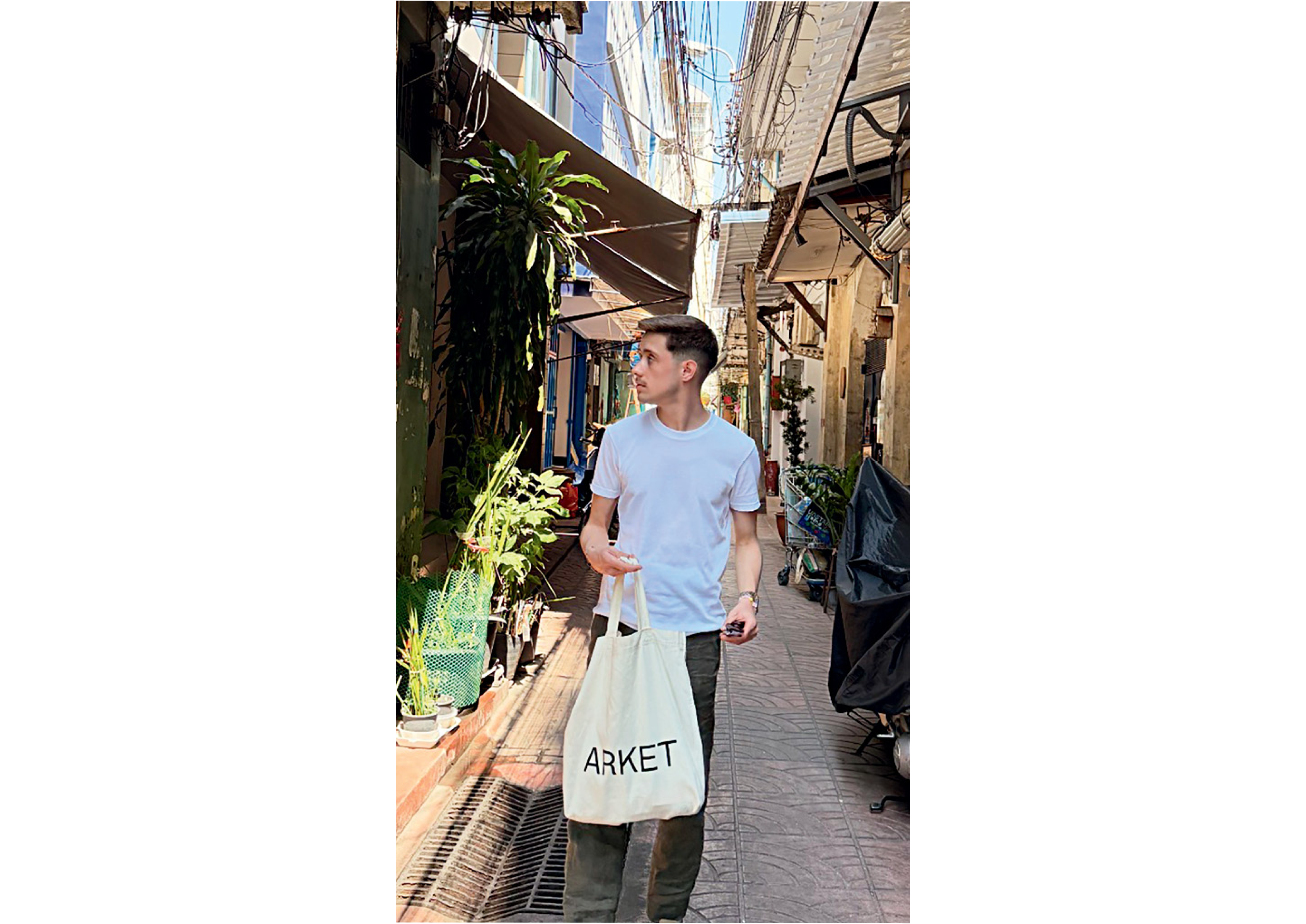 A person in a white T-shirt holds a tote bag and walks through a narrow street