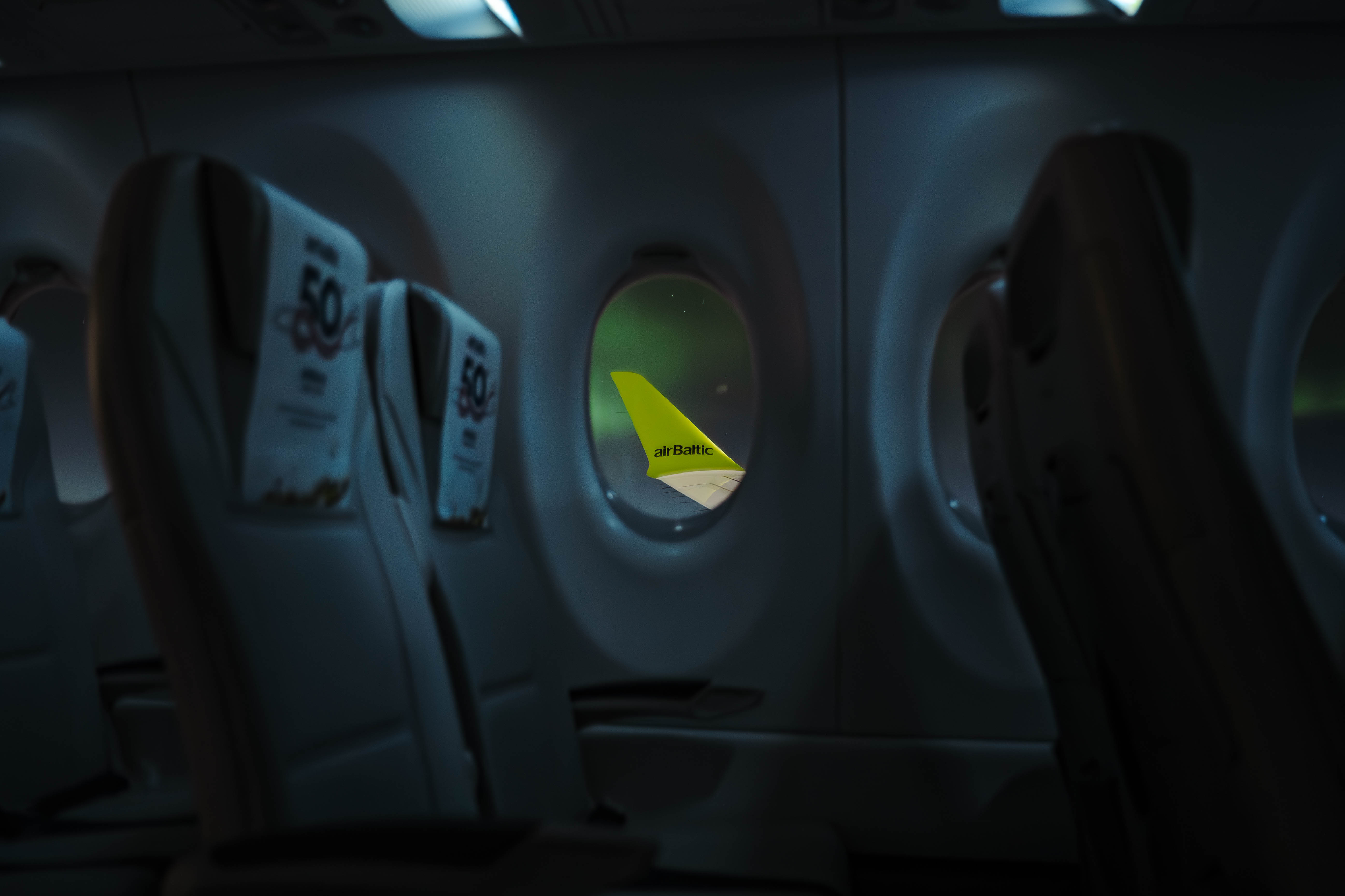 northern lights Interior of an airplane cabin with rows of seats visible. The green tail of an airBaltic plane is seen through the window.