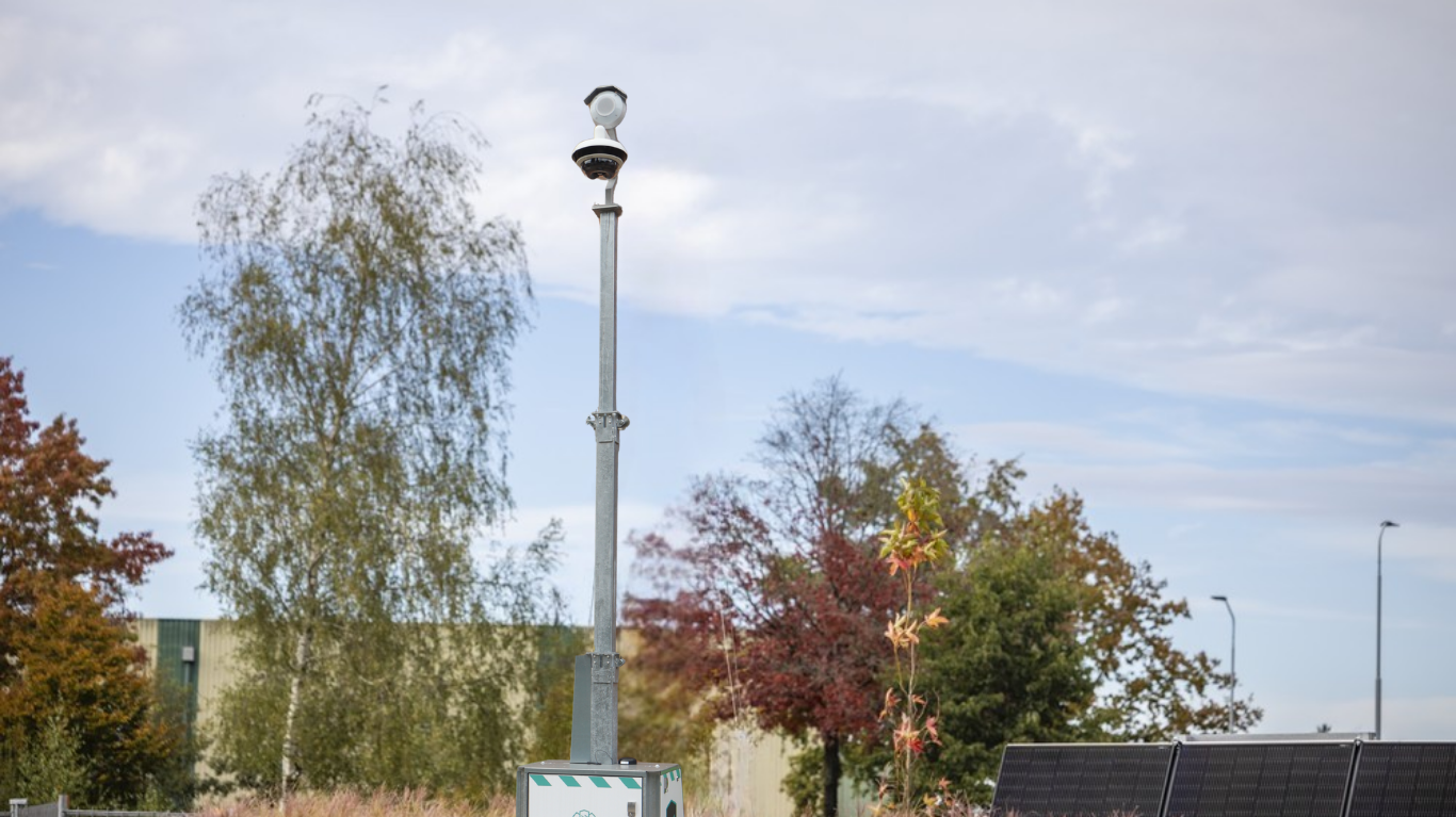 A tall surveillance camera mounted on a pole surrounded by trees and solar panels under a partly cloudy sky.