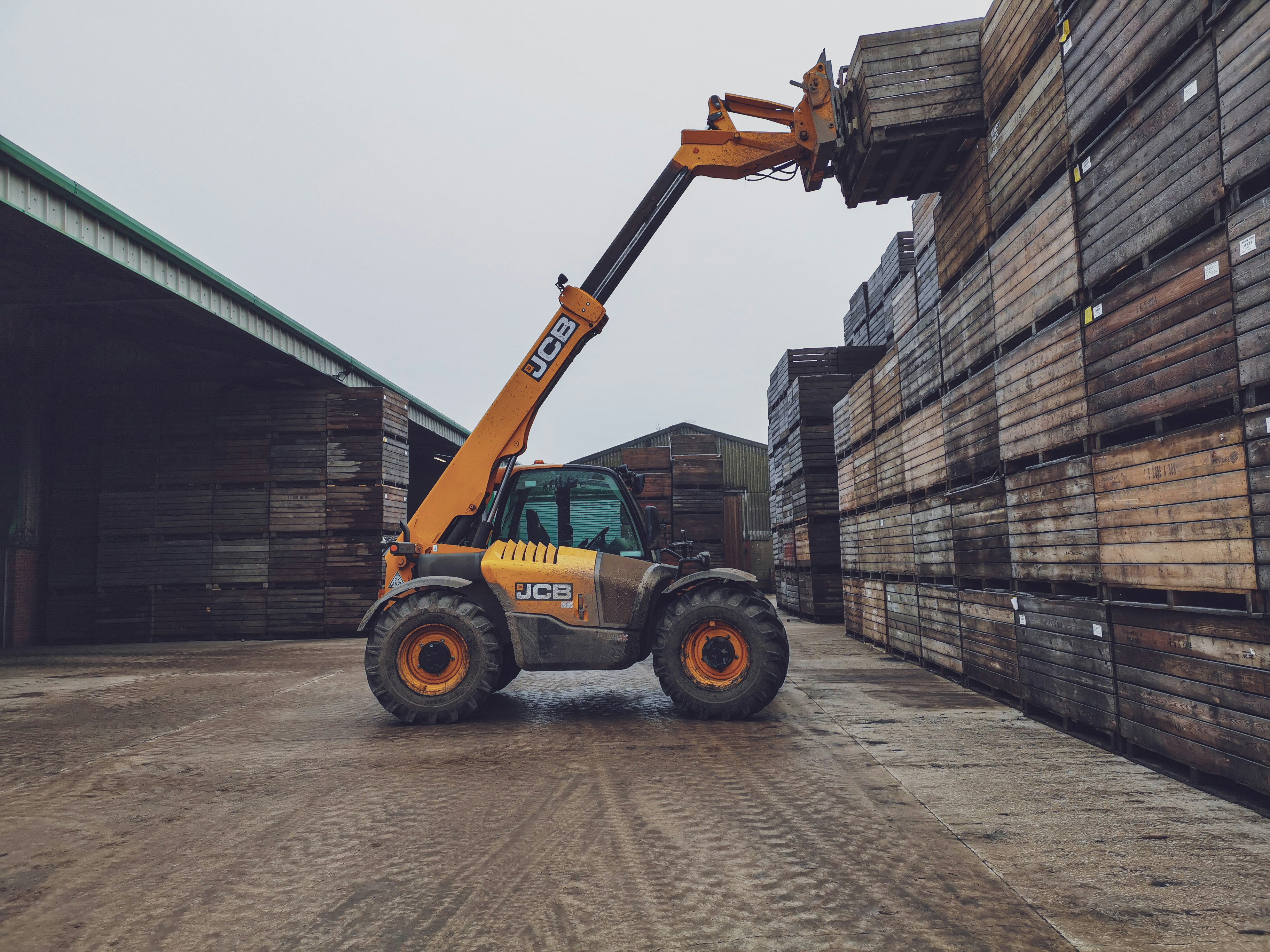 A yellow JCB telehandler lifts a wooden crate in a storage yard with stacks of crates around. Overcast sky above.