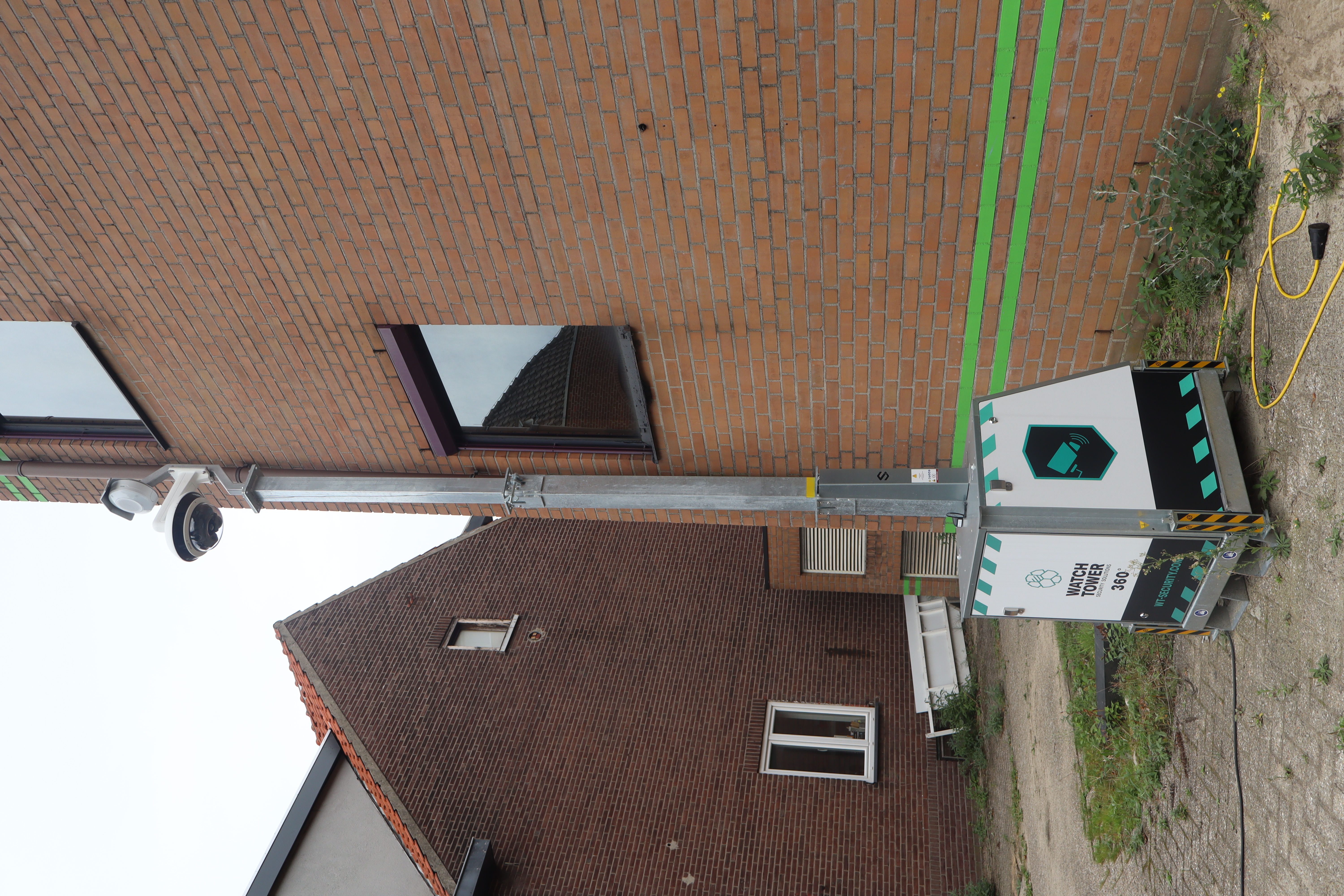 A mobile surveillance camera tower installed beside a brick building, with "Watch Tower 360°" and security logos on the base unit.