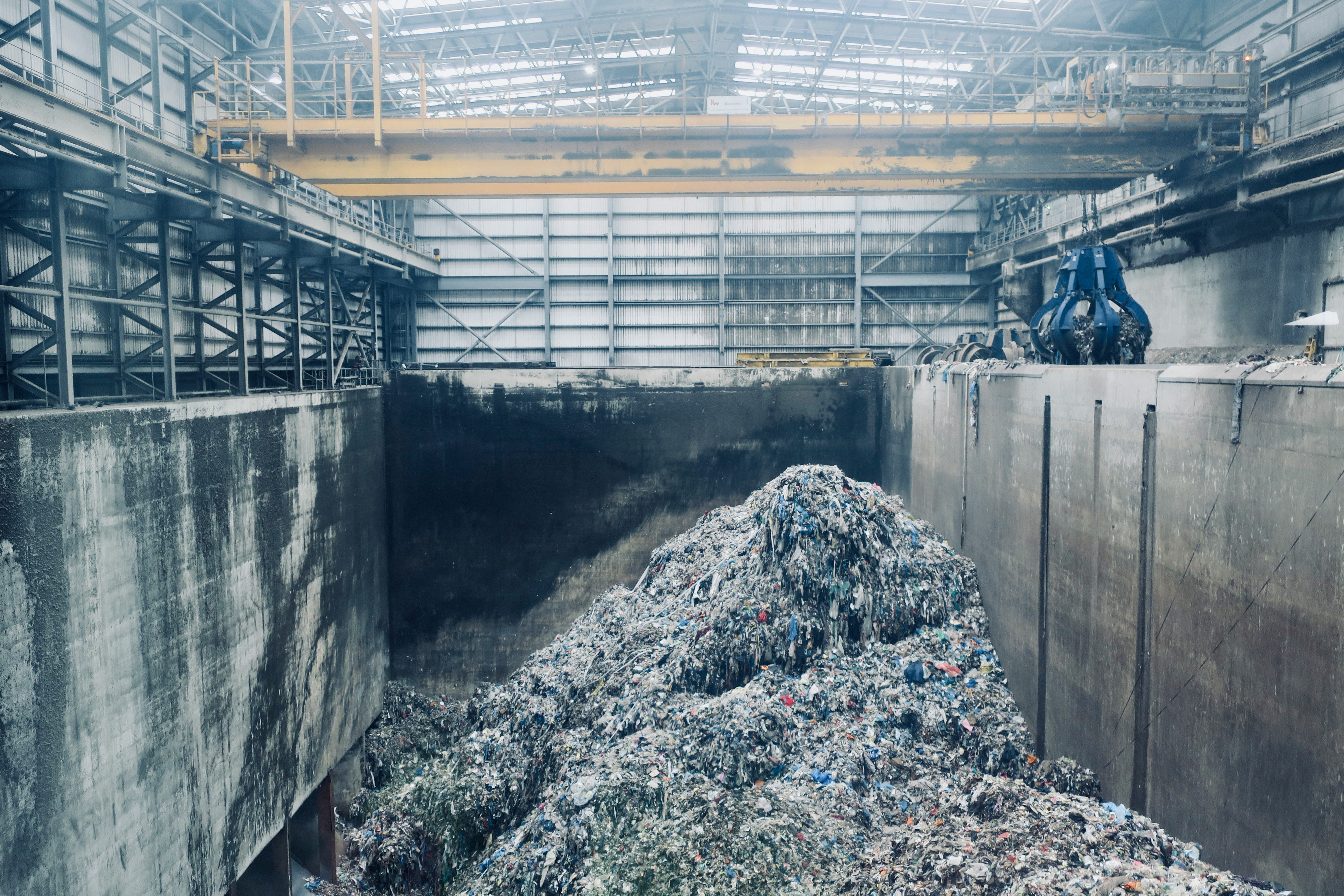 Indoor waste management facility with a large pile of mixed trash, concrete walls, steel beams, and an overhead crane with a blue grabber.