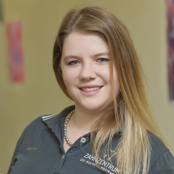 Smiling woman with long hair wearing a dark shirt stands indoors, with colorful abstract art in the blurred background.
