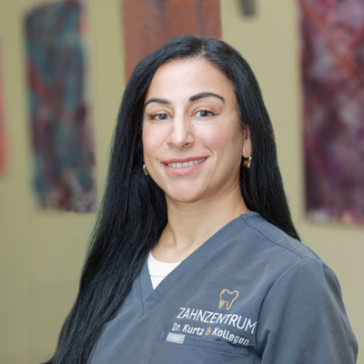 A woman with long dark hair smiles, wearing gray scrubs with a dental clinic logo, standing indoors with blurred colorful artwork behind.