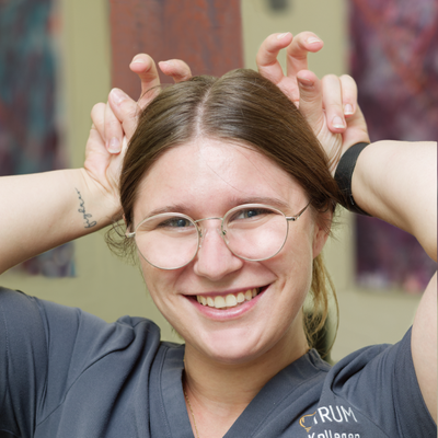 A smiling person with glasses playfully mimics animal ears using their hands, wearing a grey top with embroidered text, in a colorful room.