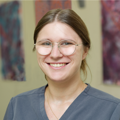 A smiling person wearing glasses and gray dental scrubs stands in front of a blurred background with colorful abstract artwork.