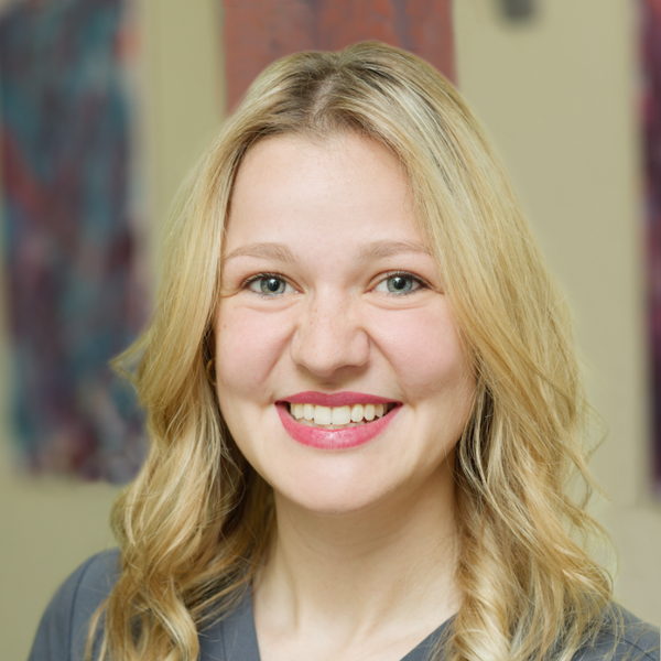 Smiling woman with blonde hair wearing a gray top, standing indoors with blurred colorful artwork in the background.