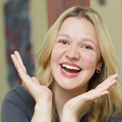 A woman with blonde hair smiling brightly, wearing a gray top, with her hands framing her face in a playful gesture.