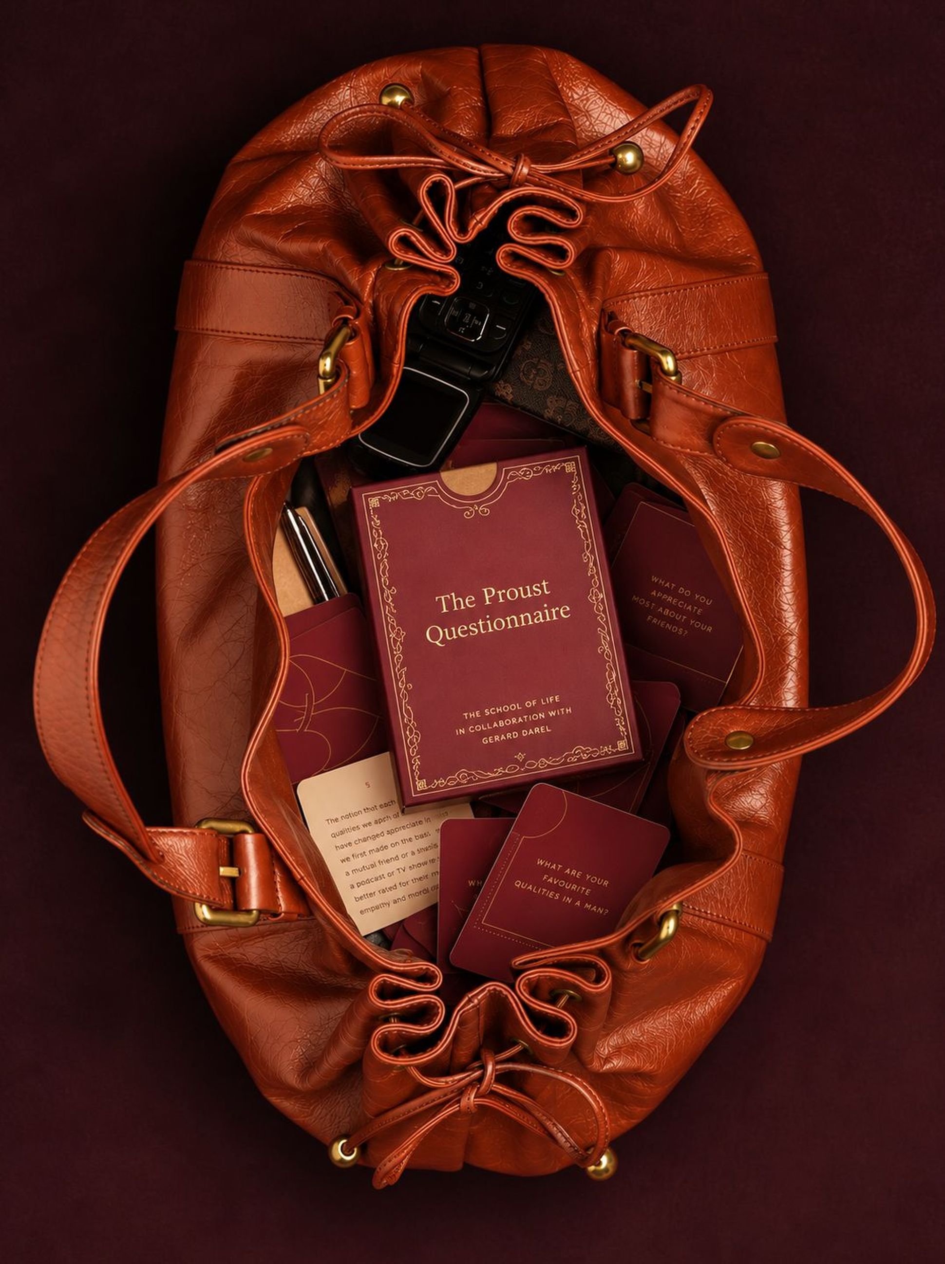 A brown leather bag containing a book titled "The Proust Questionnaire" and several matching cards, with a dark purple background.