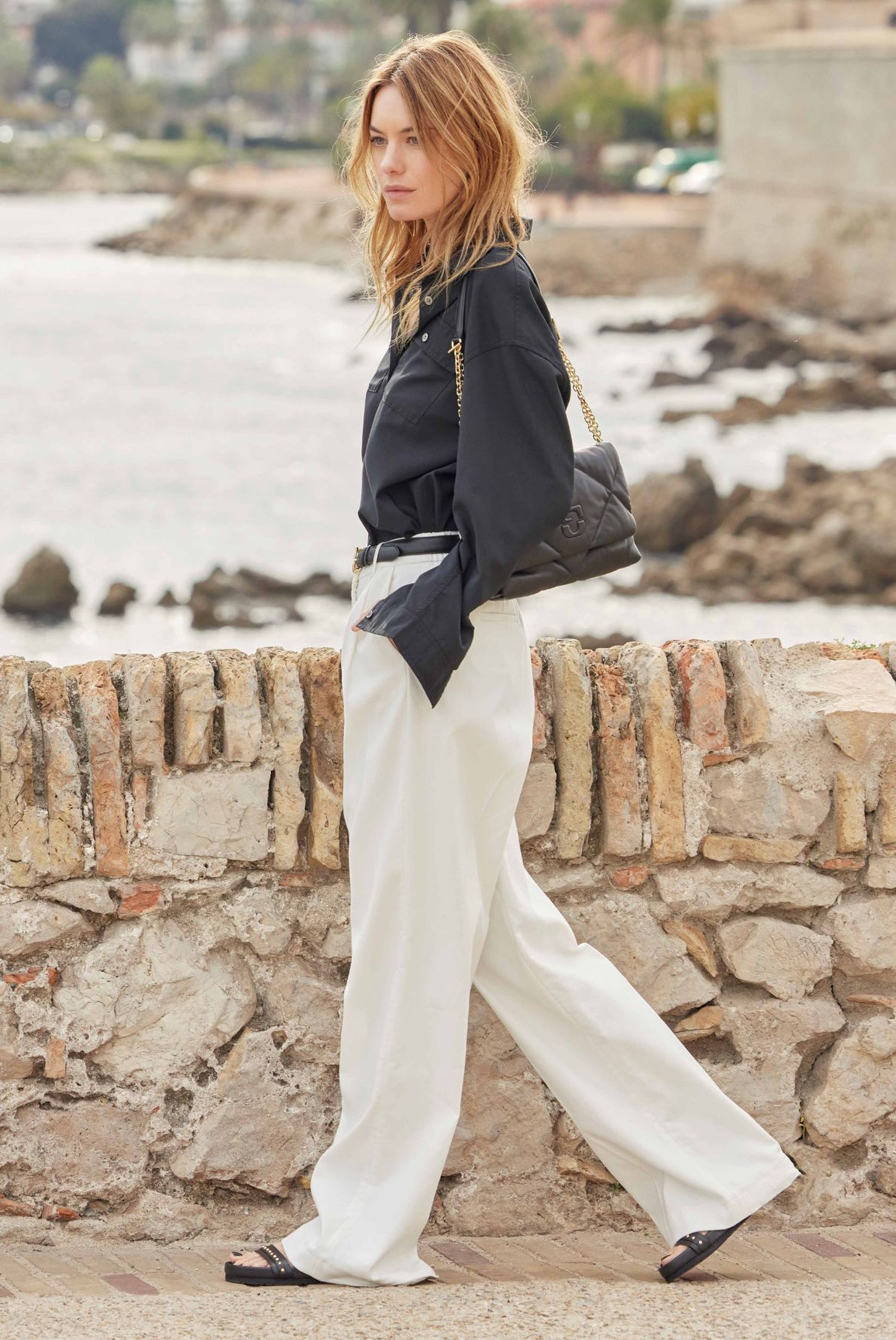 Jeune femme se promenant près de la plage, vêtue d'une chemise noire et d'un pantalon blanc