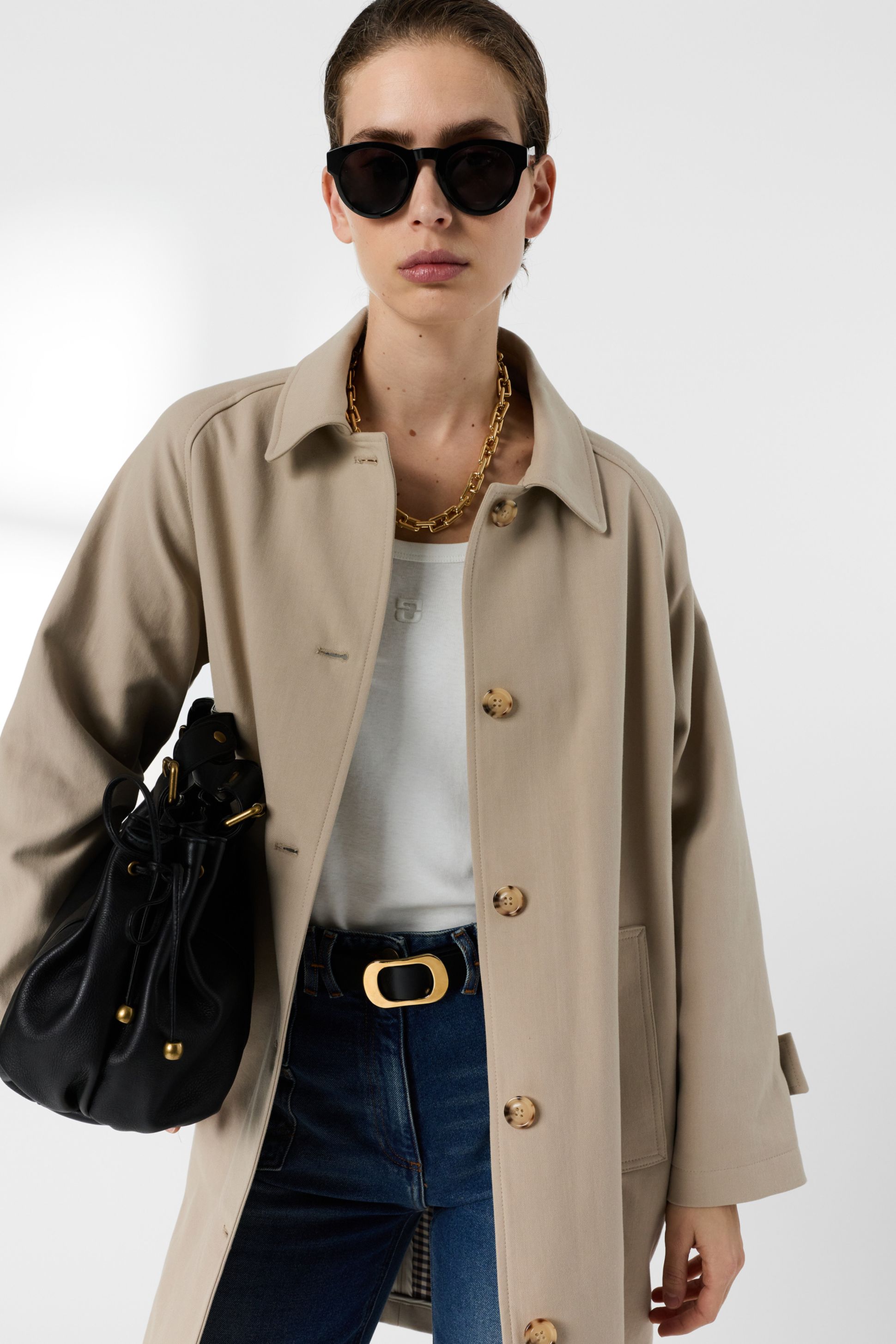 Woman wearing sunglasses, a beige coat, white shirt, and jeans, holding a black bag with gold accents, poses confidently against a plain background.