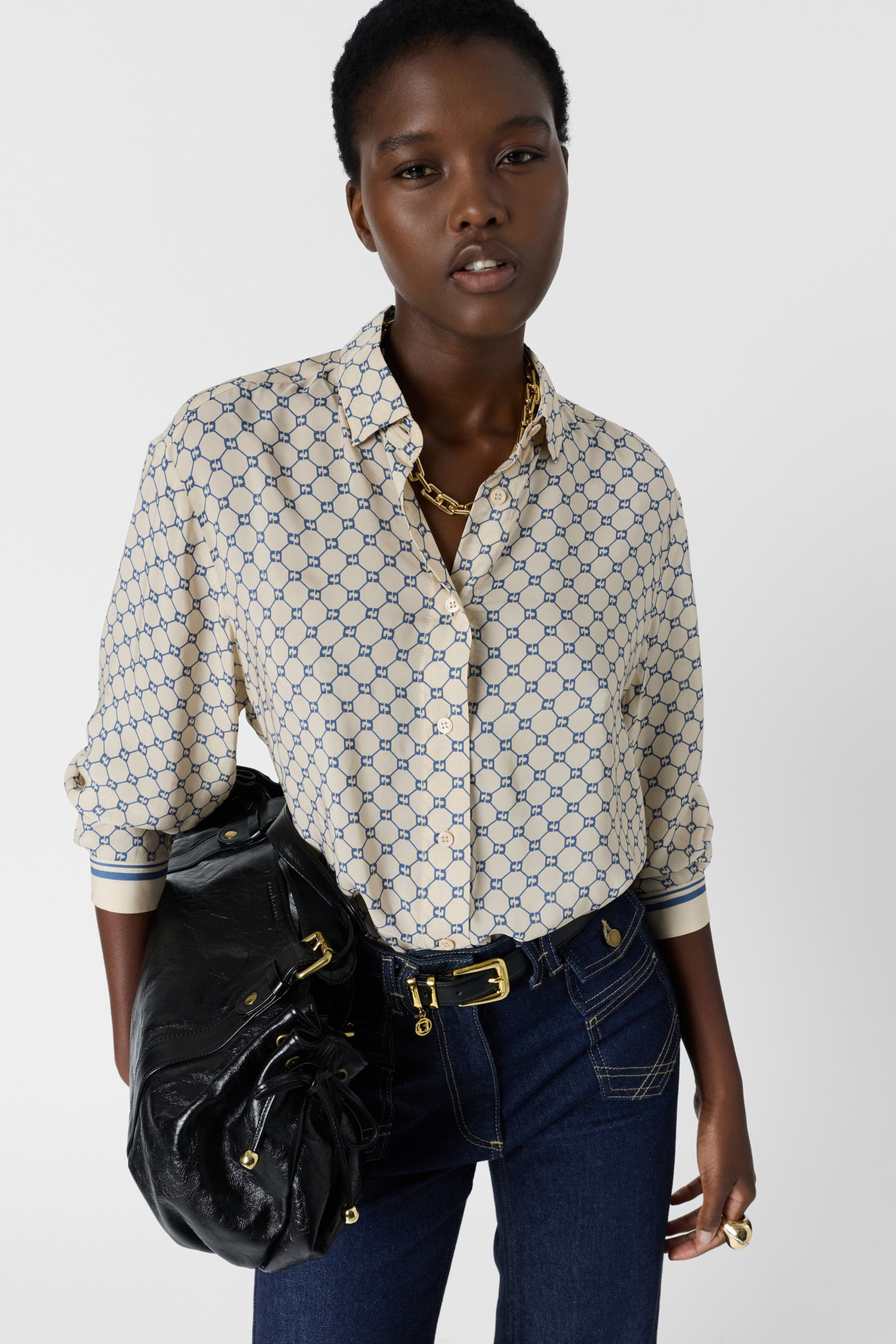 Person wearing a patterned shirt and dark jeans, holding a black bag. They have short hair and gold jewelry, standing against a plain background.