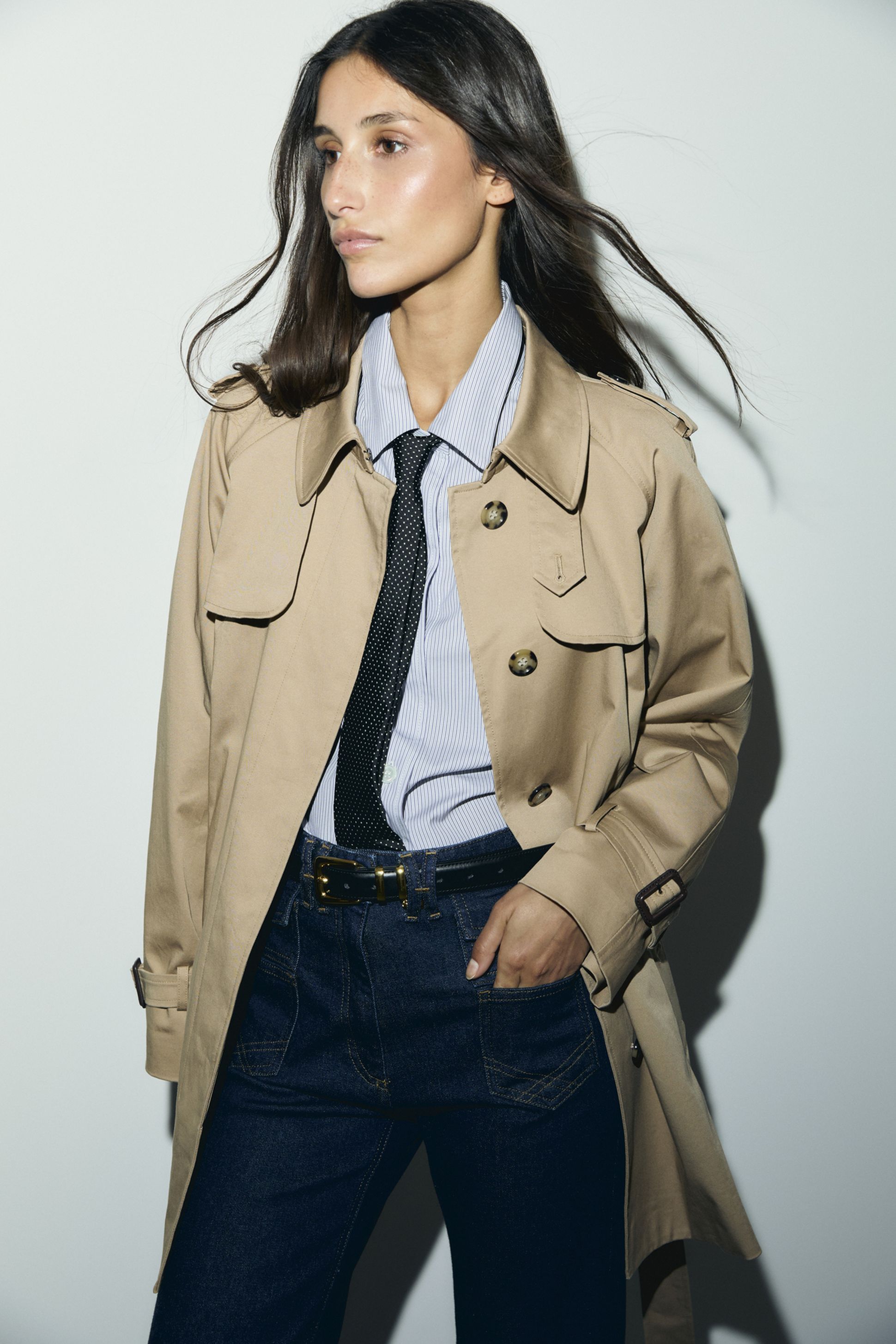A person wearing a beige trench coat over a striped shirt, black tie, and blue jeans, with long brown hair, standing against a plain background.