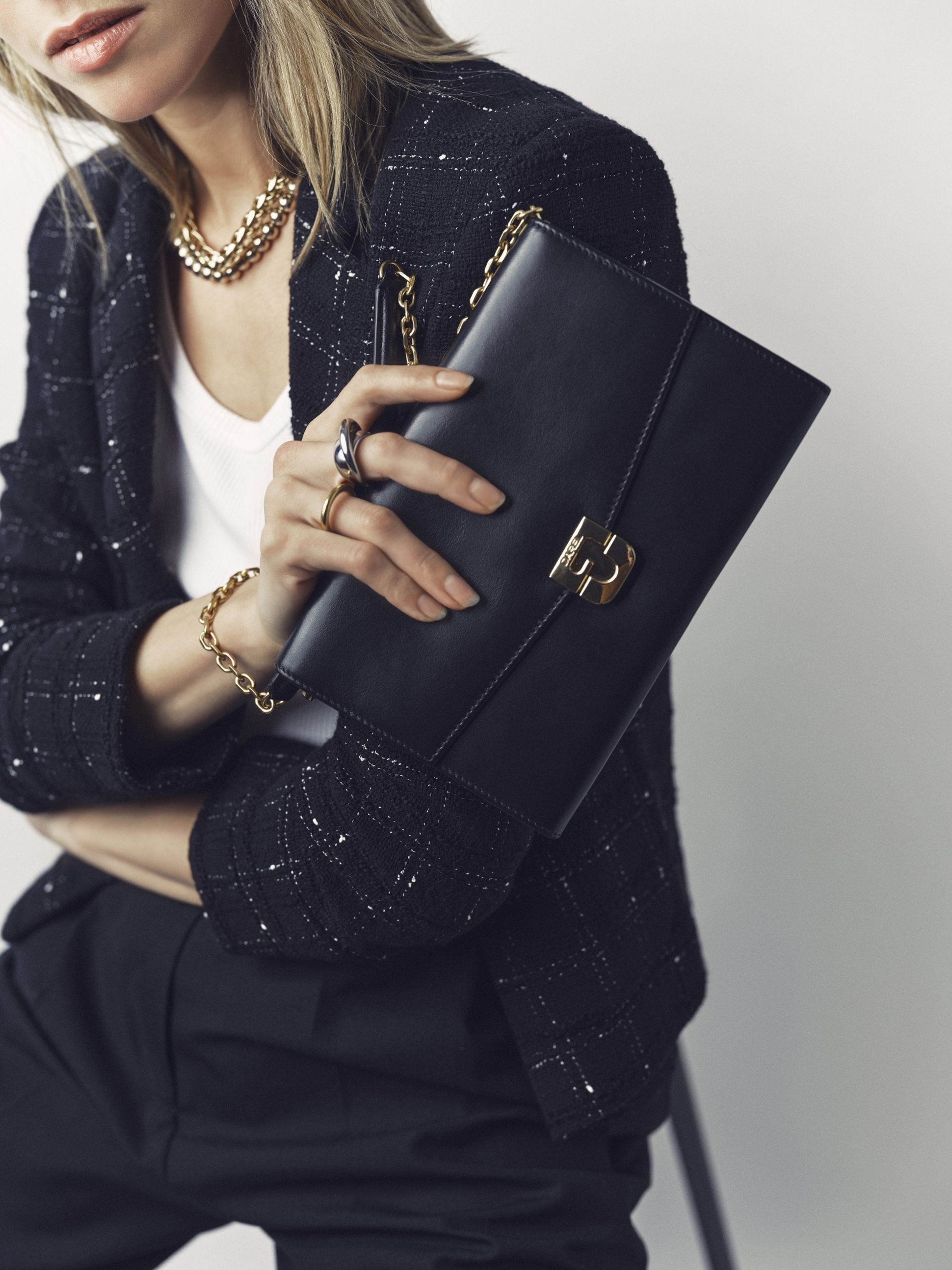 Woman holding a black clutch with a gold clasp, wearing a tweed jacket, chain necklace, and rings, sitting on a chair.