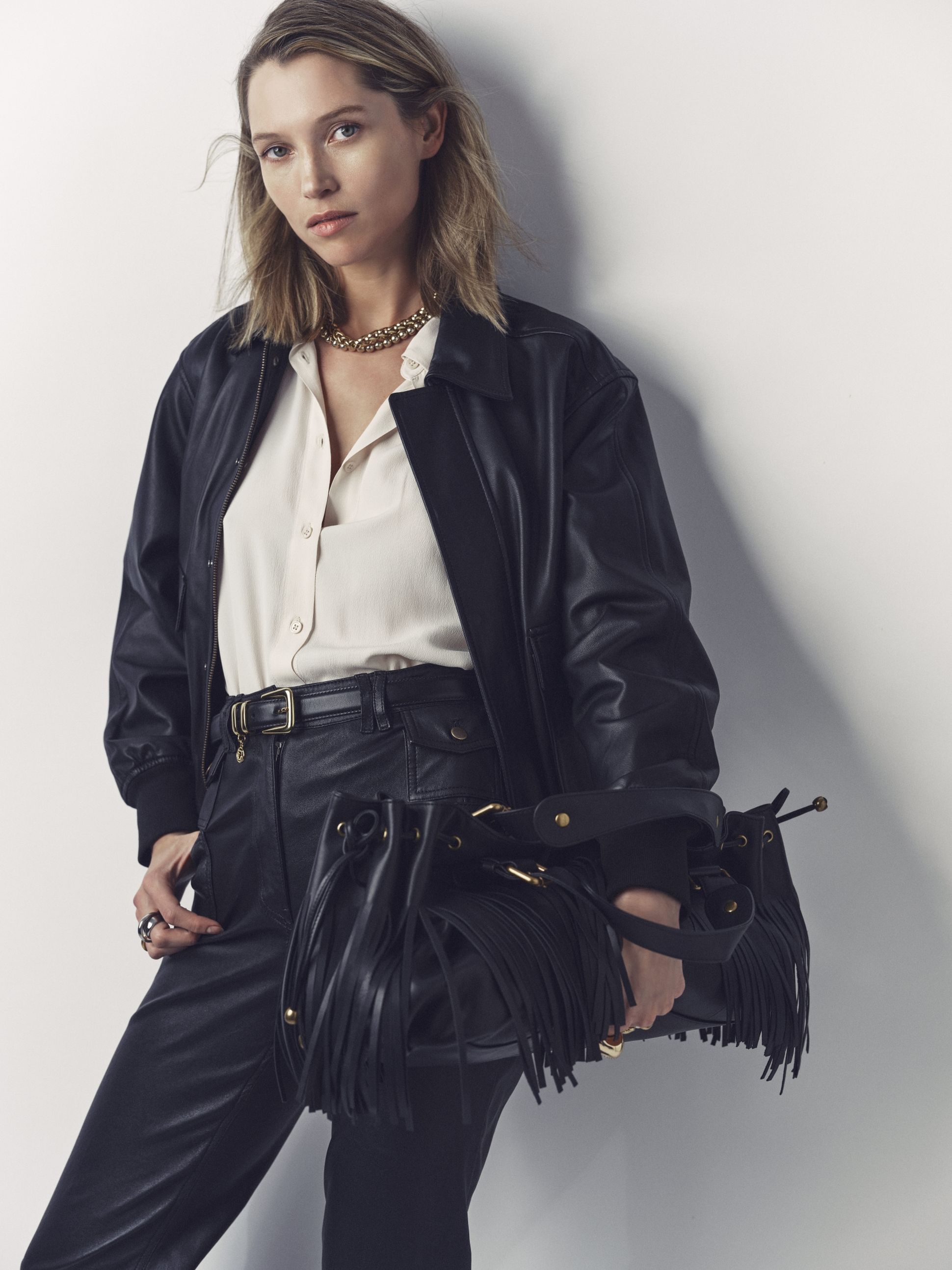 Woman in black leather outfit holding a fringed handbag, wearing a white shirt and gold necklace, standing against a plain background.