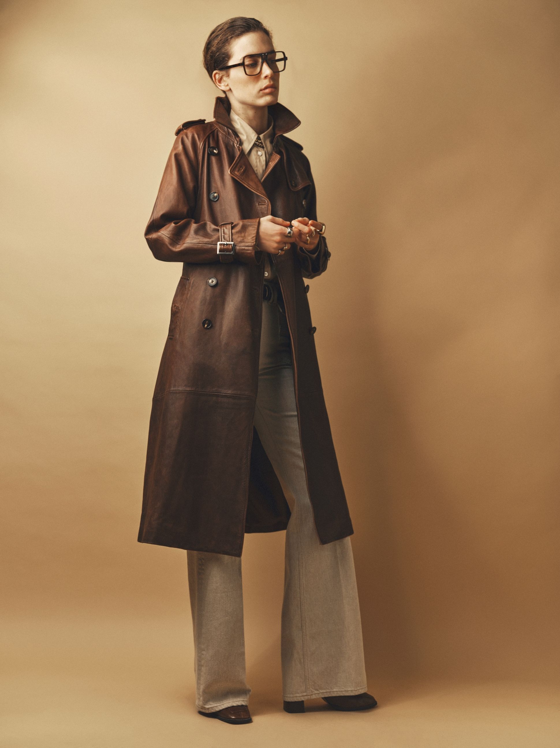 Person wearing a long brown leather coat, glasses, and flared pants, standing against a beige background, holding an object in hand.