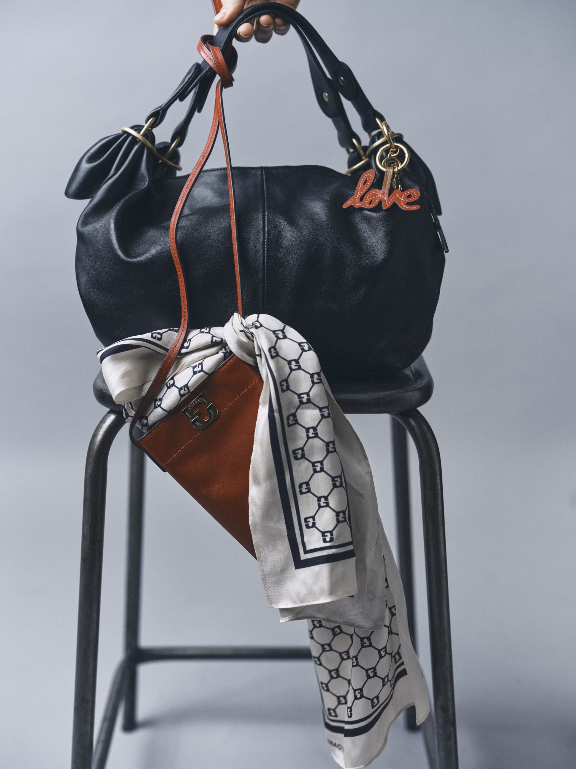 A black handbag with "love" charm hangs on a stool, featuring a draped white-and-blue patterned scarf and a brown leather pouch.