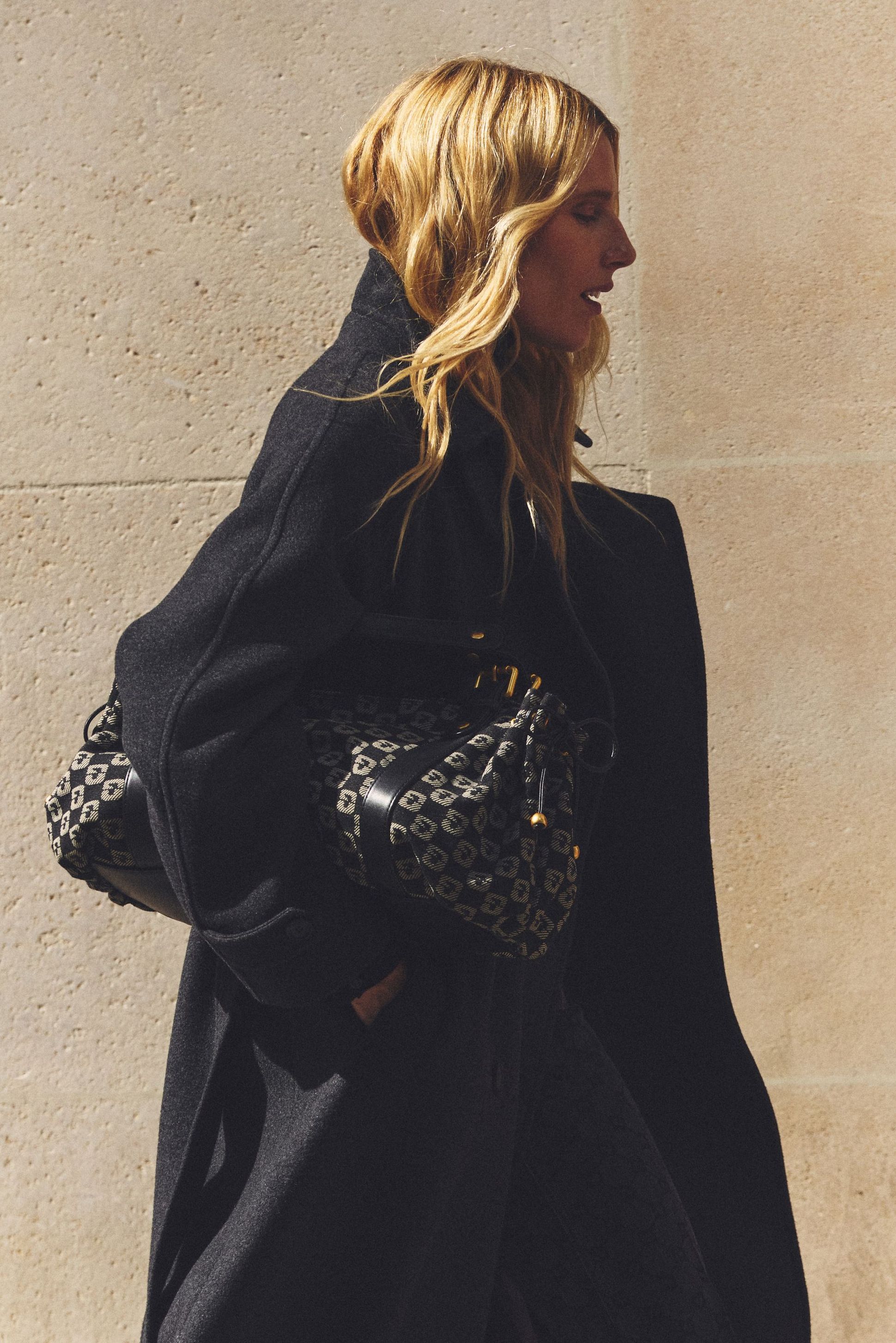 A woman with wavy blonde hair wearing a black coat carries a patterned handbag, walking past a textured beige wall.