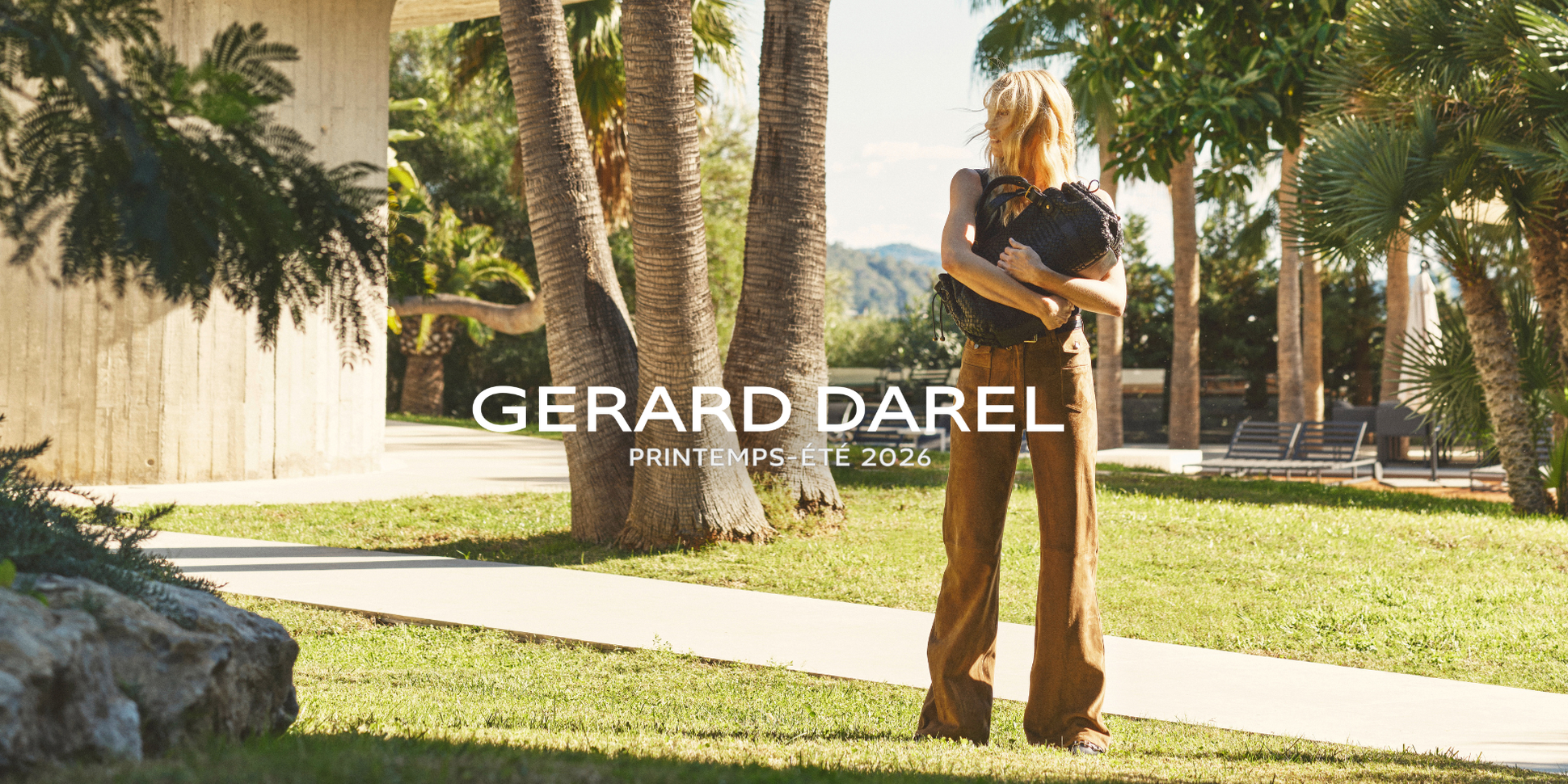 Woman in brown outfit holding handbag, standing outdoors near palm trees and a modern building, with "Gerard Darel Printemps-Été 2026" text.