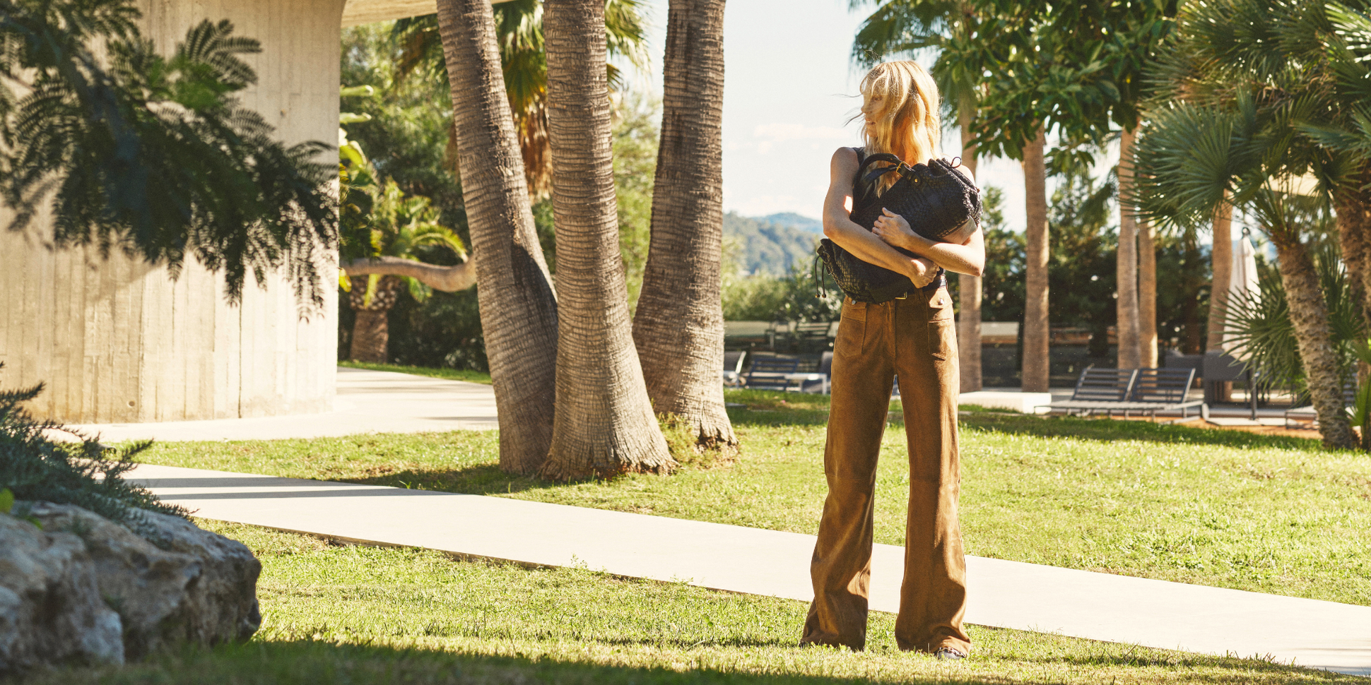 Woman in brown pants and black top stands in a sunlit garden, holding a small black dog. Palm trees and a concrete building are in the background.