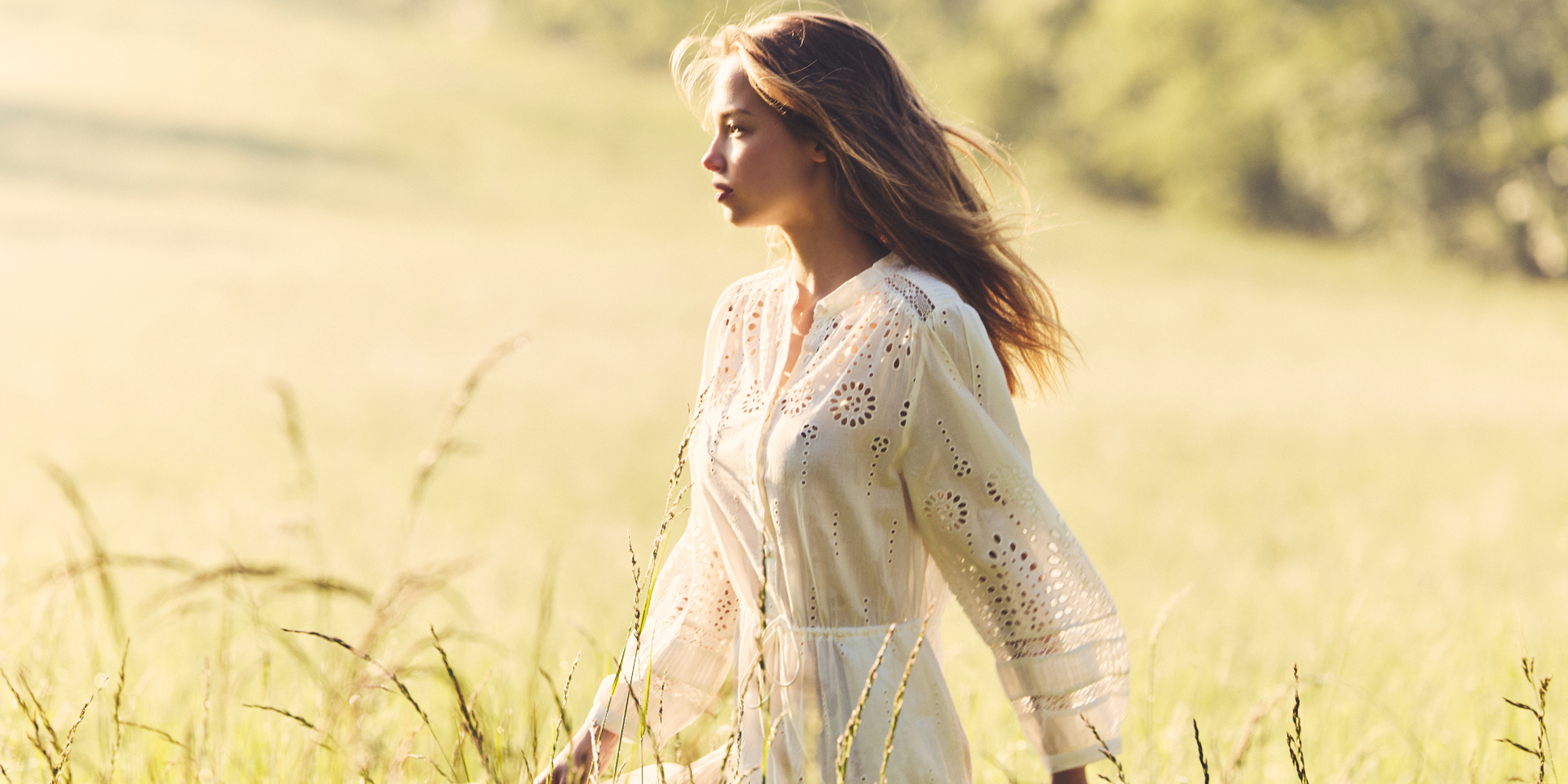 Woman in a white eyelet dress walks through a sunlit grassy field, with gentle light and a soft background.