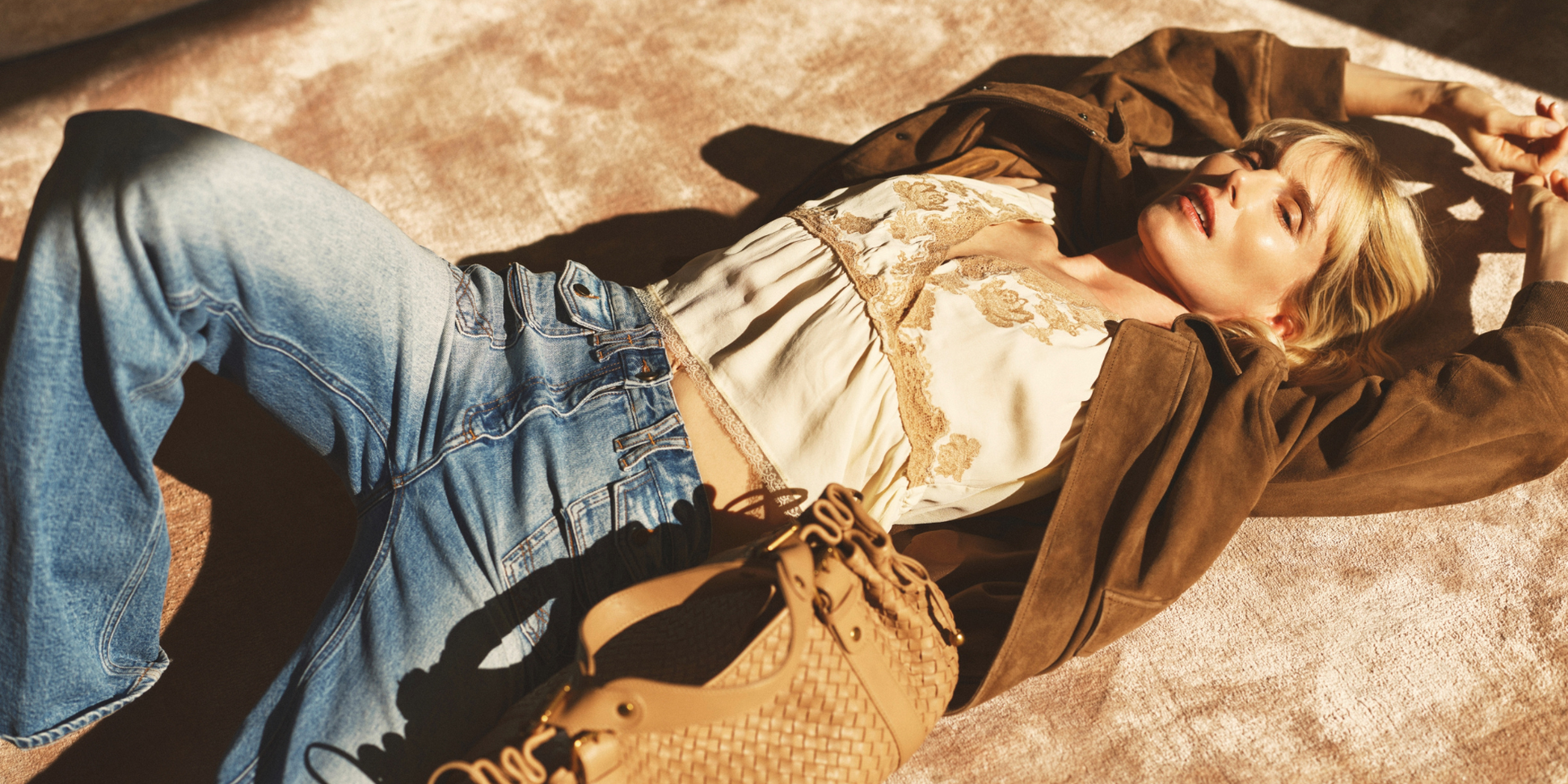 Person lying on a carpet in sunlight, wearing jeans, a lace top, and a brown jacket, with a woven beige bag beside them.