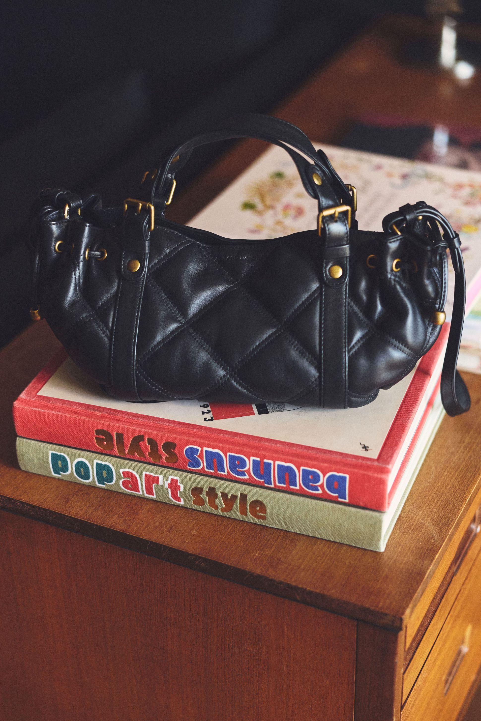 A black quilted handbag with gold hardware rests on a stack of two colorful books on a wooden tabletop.
