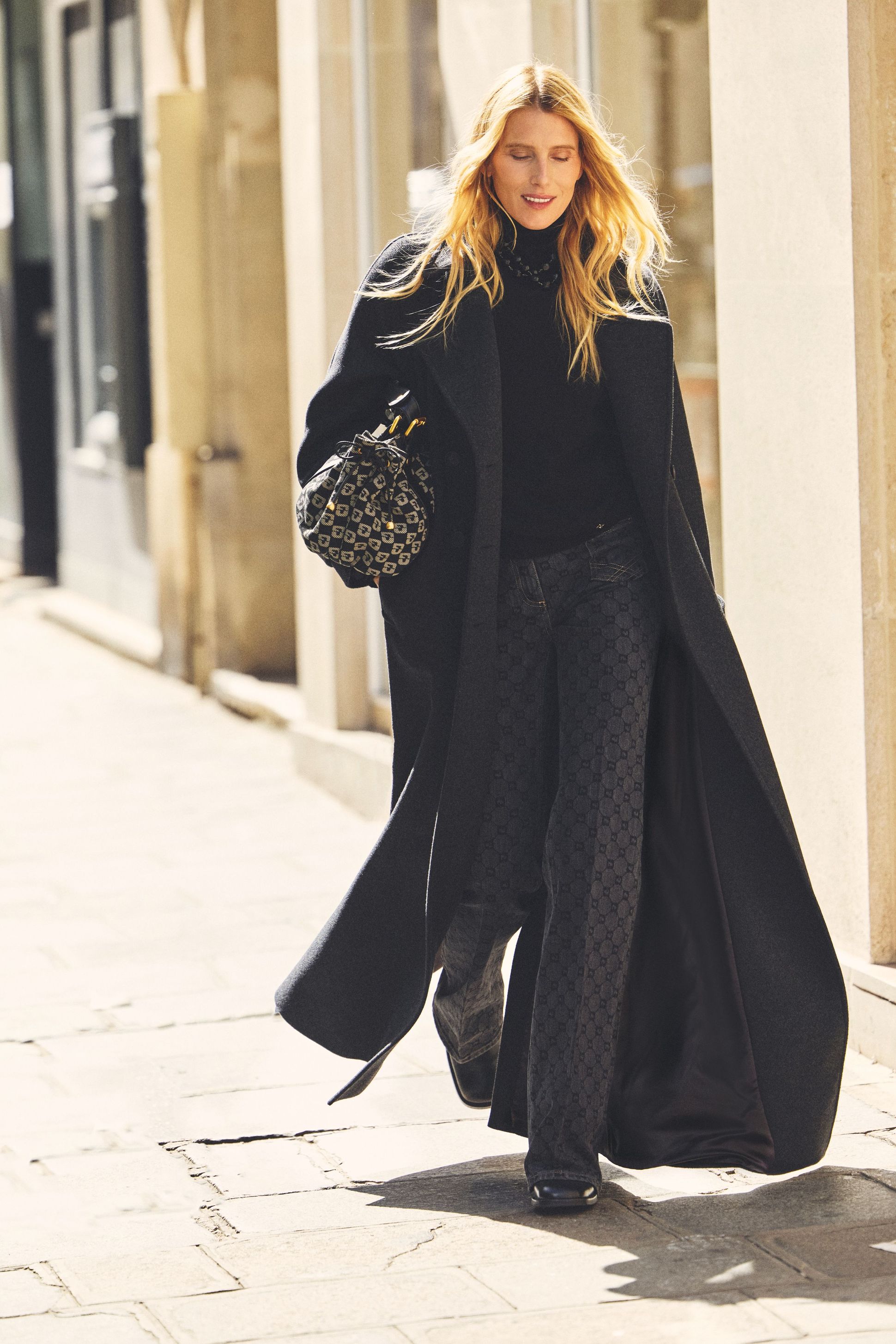 Woman in a long black coat and patterned pants walking on a sunlit street, carrying a patterned bag, with blonde hair flowing.