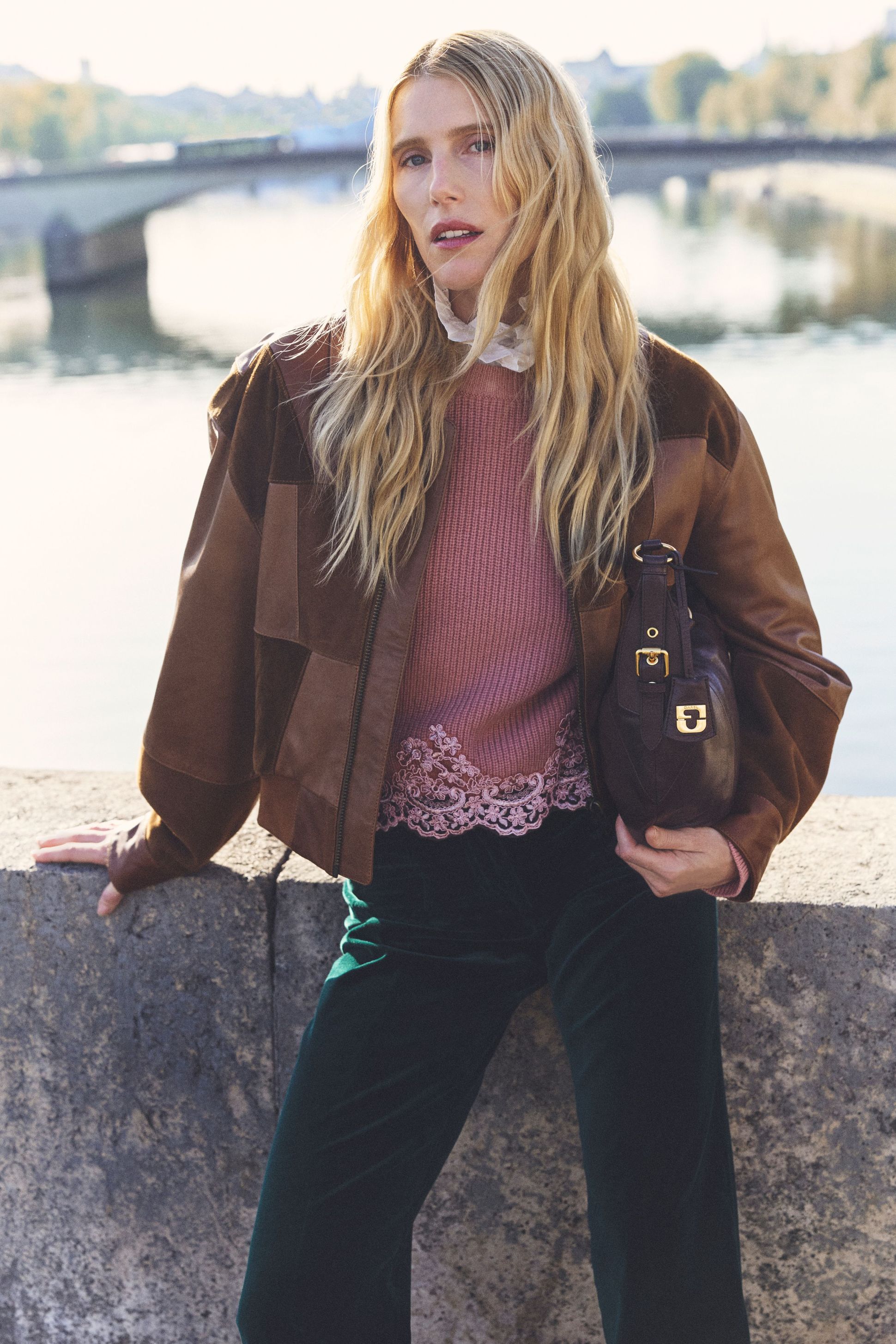 Woman with long blonde hair in a brown jacket and pink top leaning against a stone railing, with a river and bridge in the background.