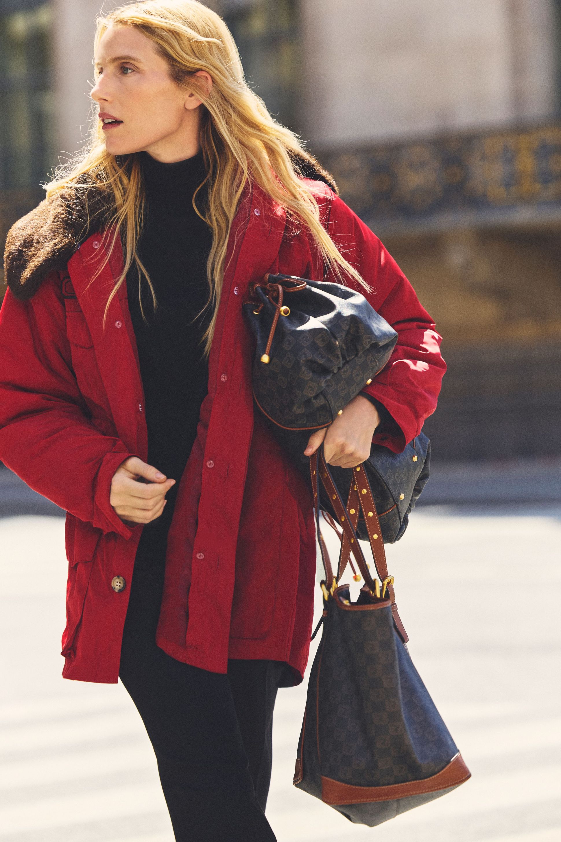 Woman in a red coat, holding handbags, looks to the side while walking on a sunny street.