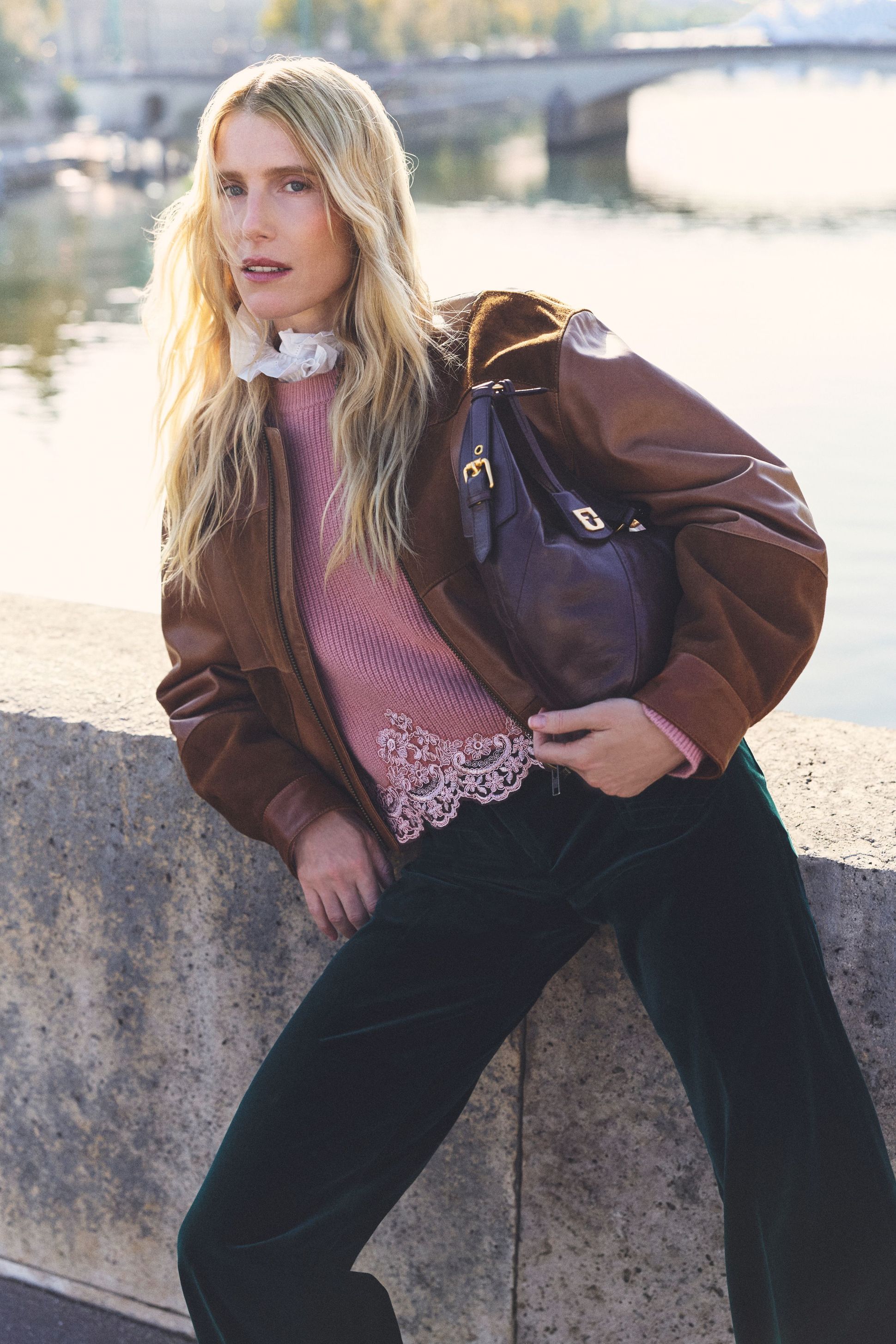 A person with long blonde hair leans against a stone railing by a river, wearing a brown jacket, pink sweater, and velvet pants.