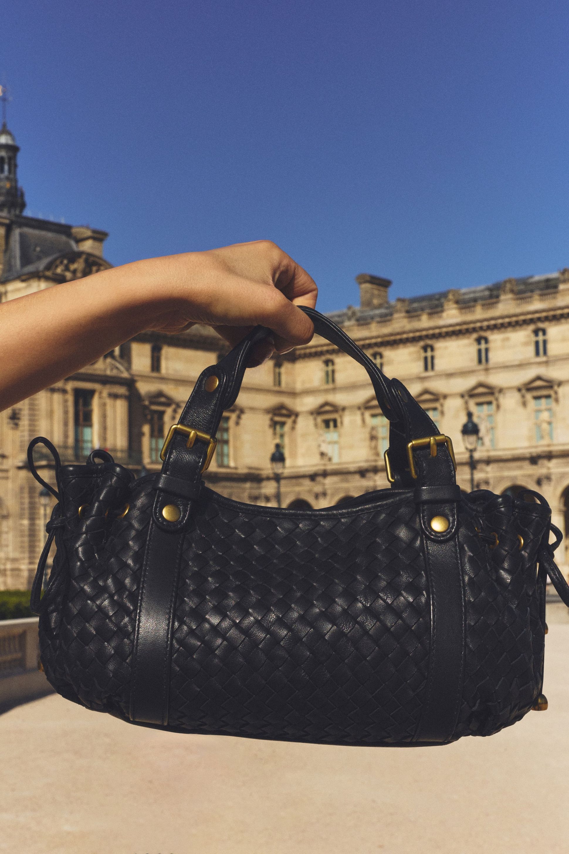A hand holding a woven black leather handbag with brass accents, set against a historic building under a clear blue sky.