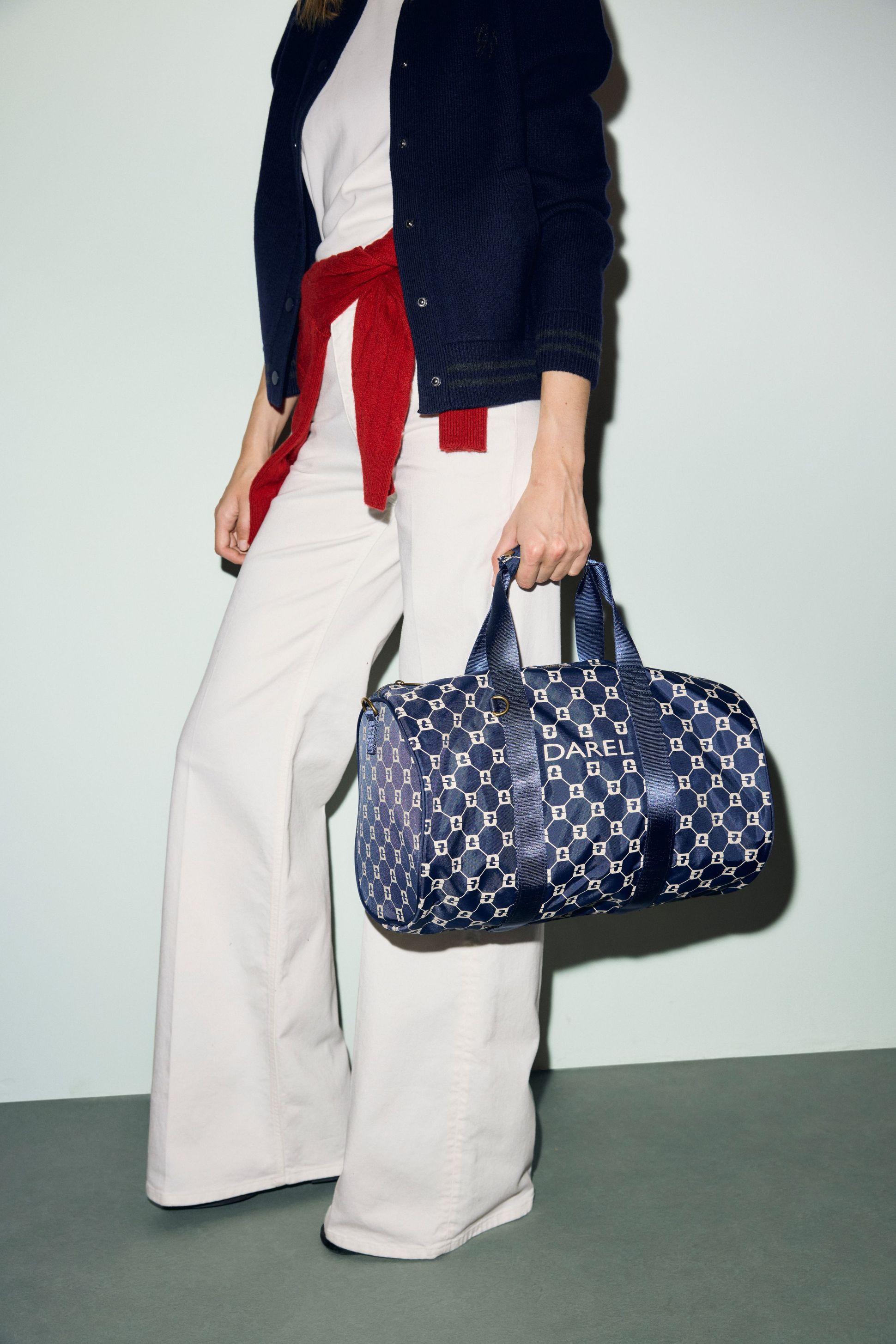 Person wearing white pants and a navy jacket, holding a blue patterned bag with "DAREL" printed on it. A red sweater is tied at the waist.