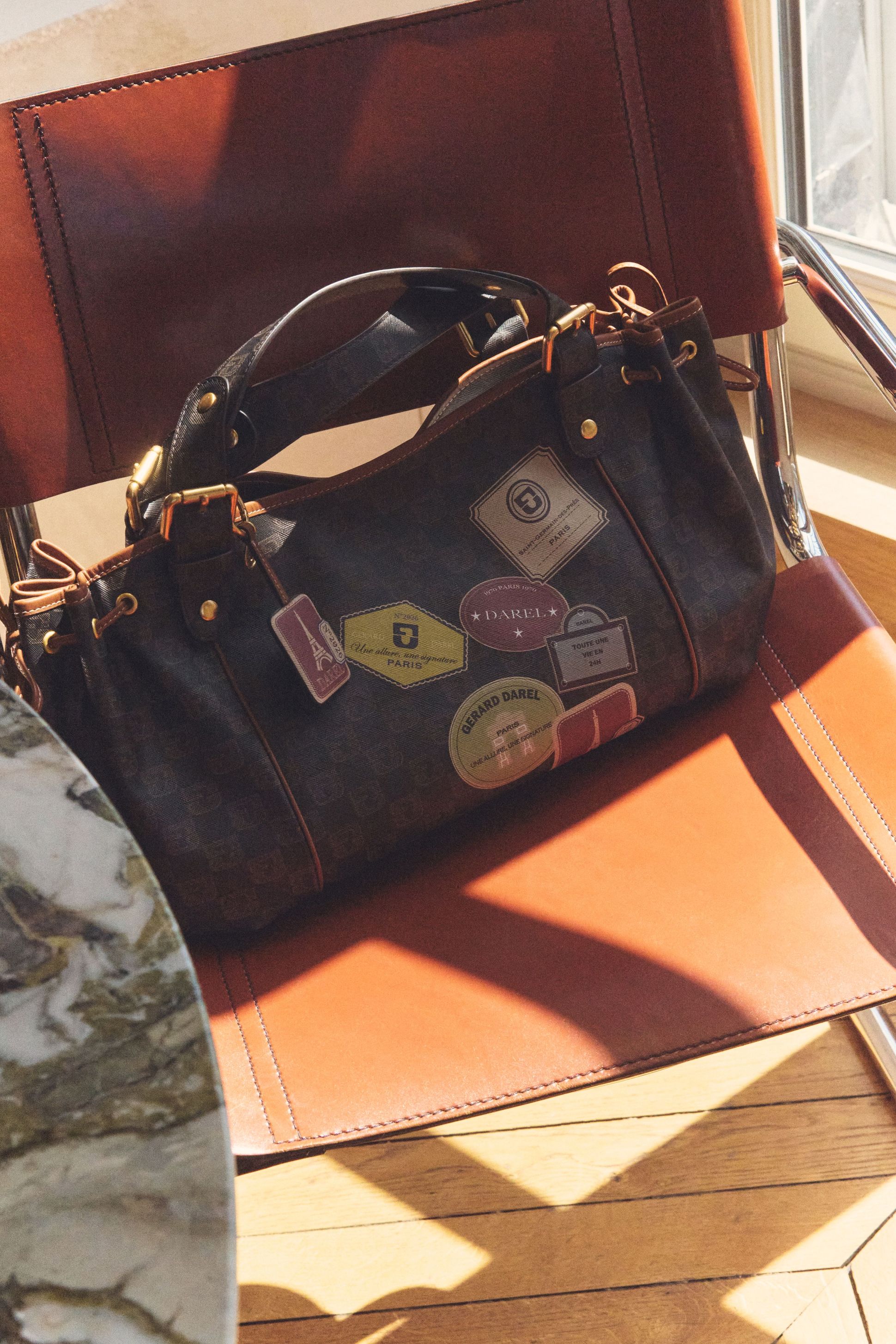 A brown handbag with travel-themed patches sits on a brown leather chair under sunlight, with a marble table in view.