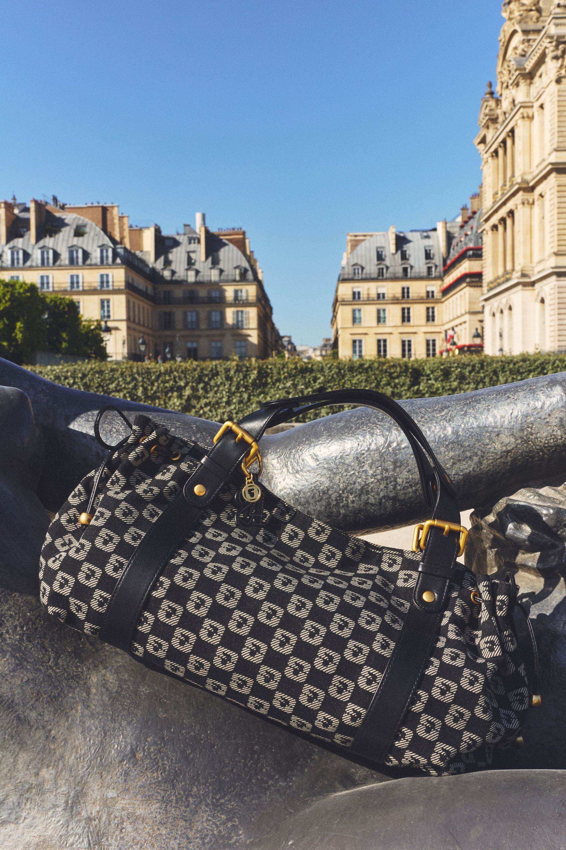 Patterned handbag with leather straps on a statue, against a backdrop of historic buildings and a clear blue sky.